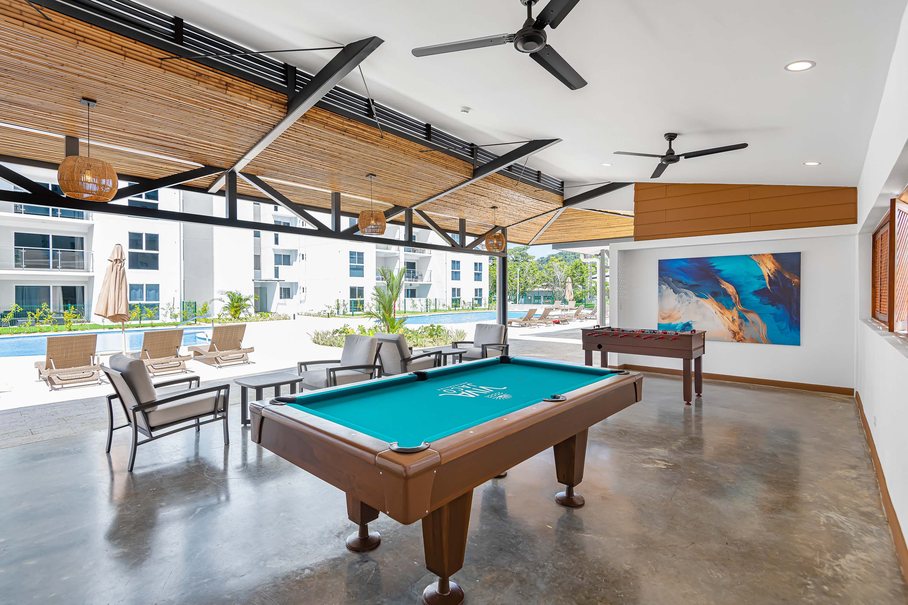Open-air game room with a pool table and foosball table under a high ceiling with exposed beams and bamboo accents. Comfortable seating areas are arranged around the space, which overlooks the outdoor pool area.