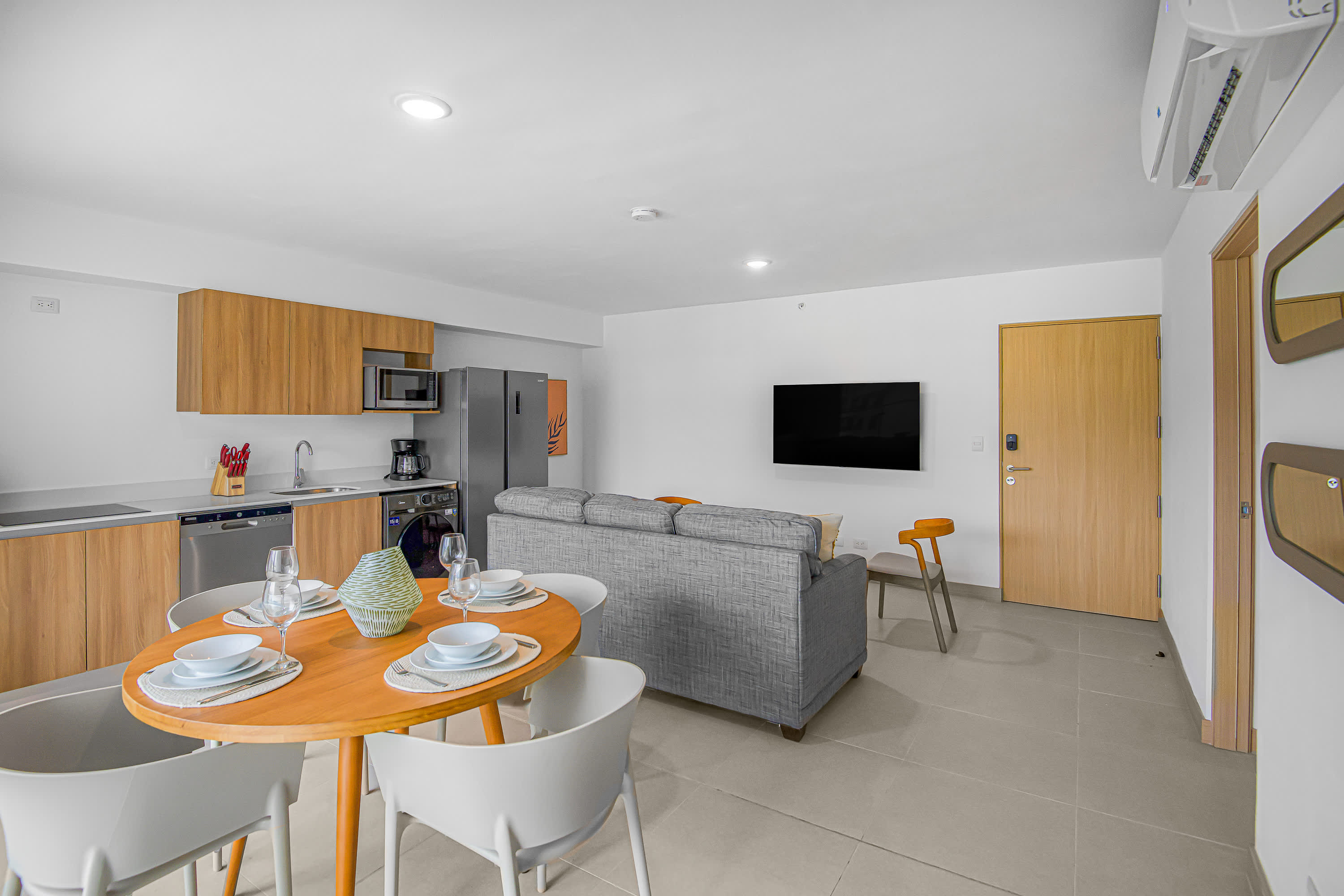 Modern open-concept living space featuring a gray sofa, a wooden dining table with white chairs, and a compact kitchen with light wood cabinetry and stainless steel appliances. A wall-mounted TV and an air conditioning unit are visible.