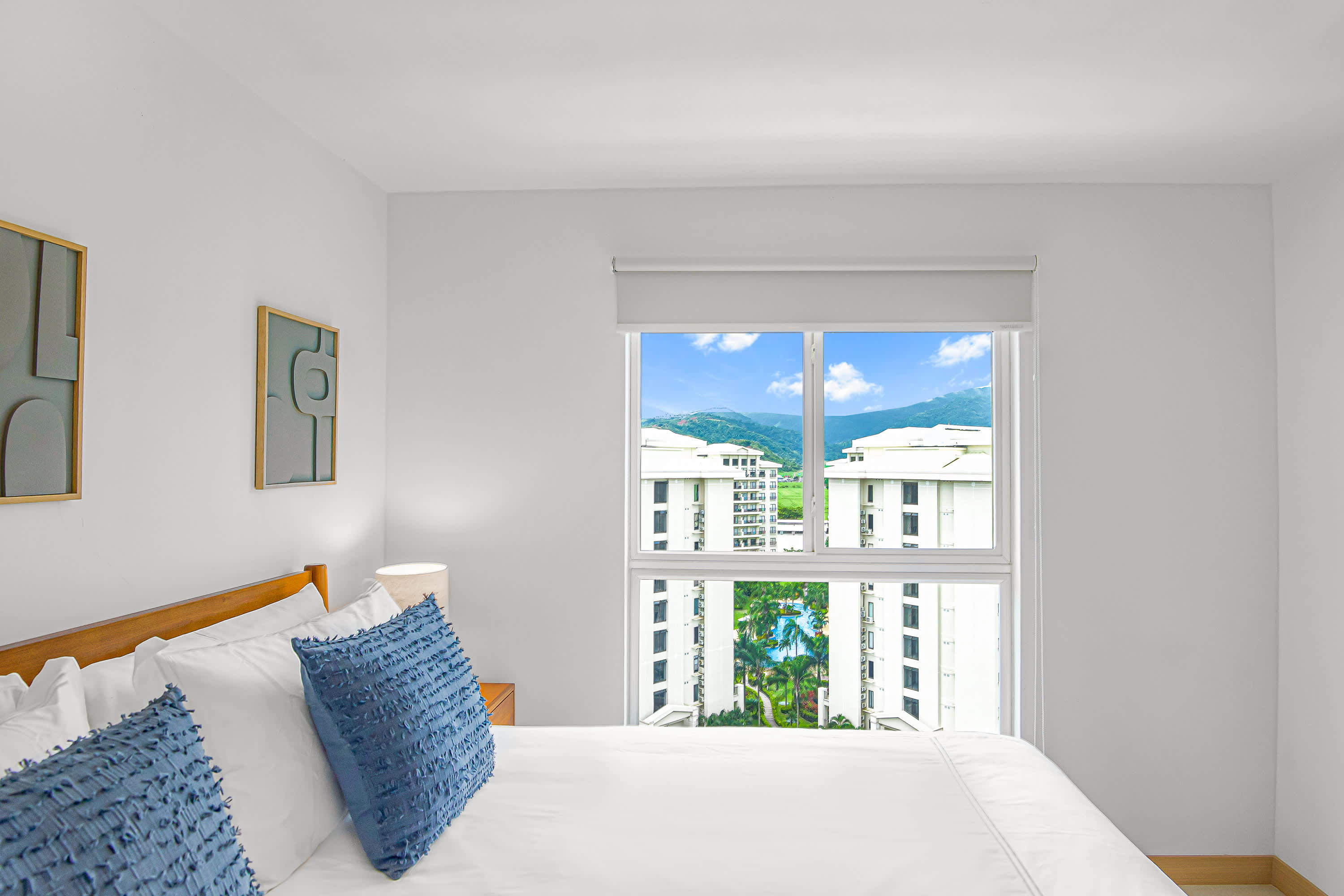 Bedroom with a large window offering a view of other buildings, a swimming pool, and distant mountains. The bed is dressed in white linens with blue accent pillows, and abstract art adorns the wall.