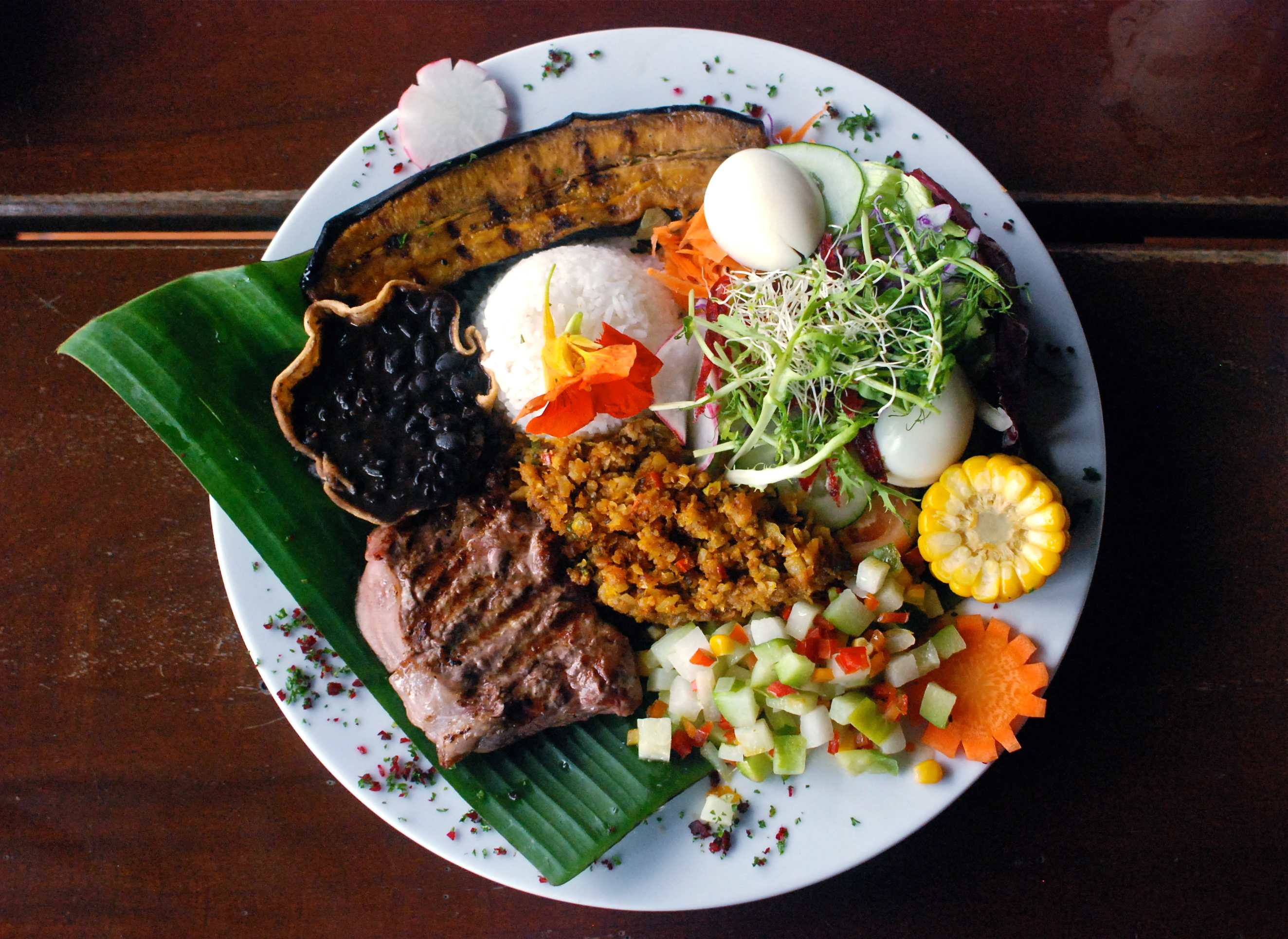 A vibrant and generously portioned meal served on a white plate, featuring grilled meat, rice, black beans in a plantain cup, a hard-boiled egg, corn, and a fresh salad with various vegetables and sprouts. The dish is garnished with a banana leaf and edible flowers.