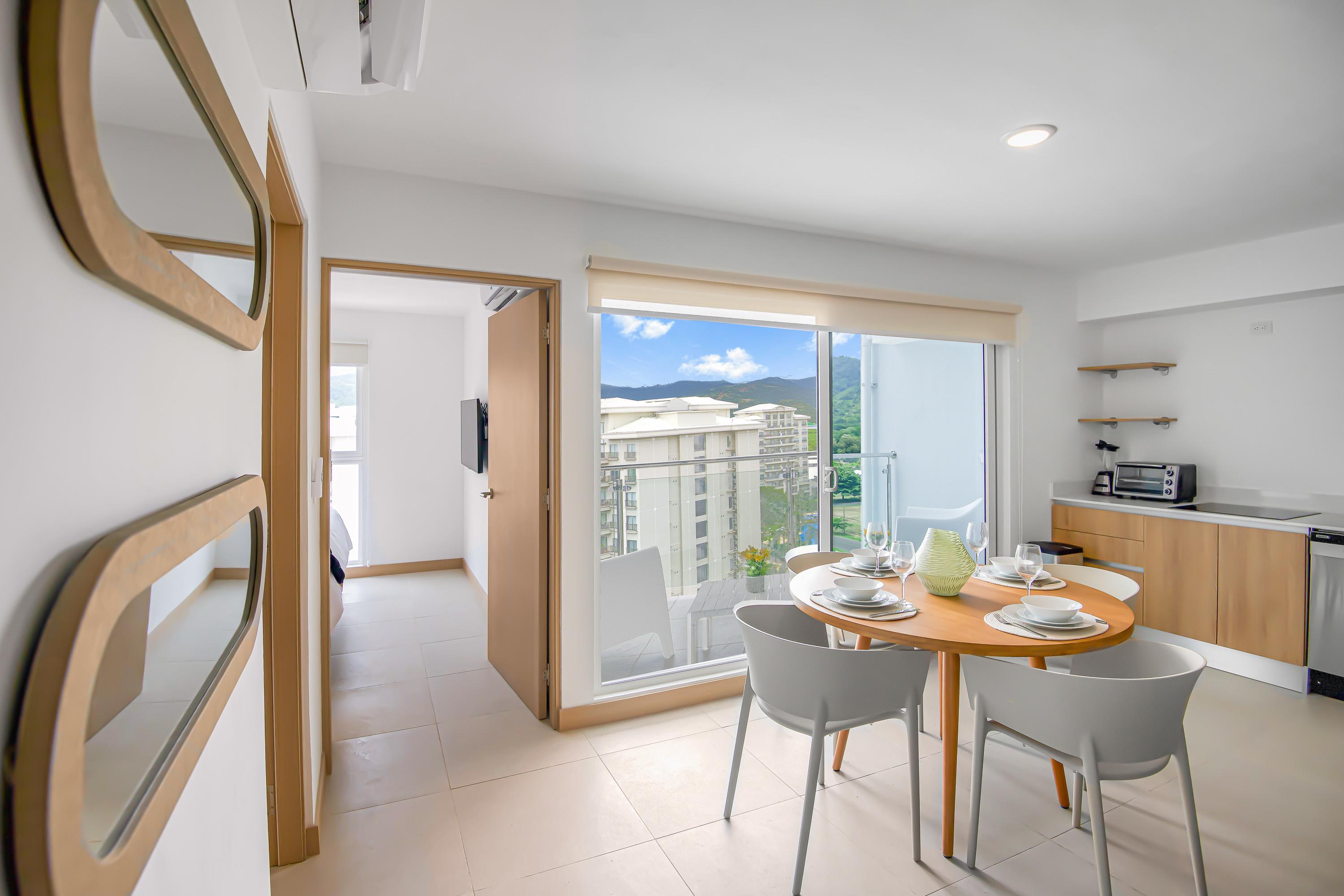 Bright dining area with a round wooden table and white chairs, adjacent to a living space with a gray sofa. Large sliding glass doors open to a balcony offering views of buildings and mountains.