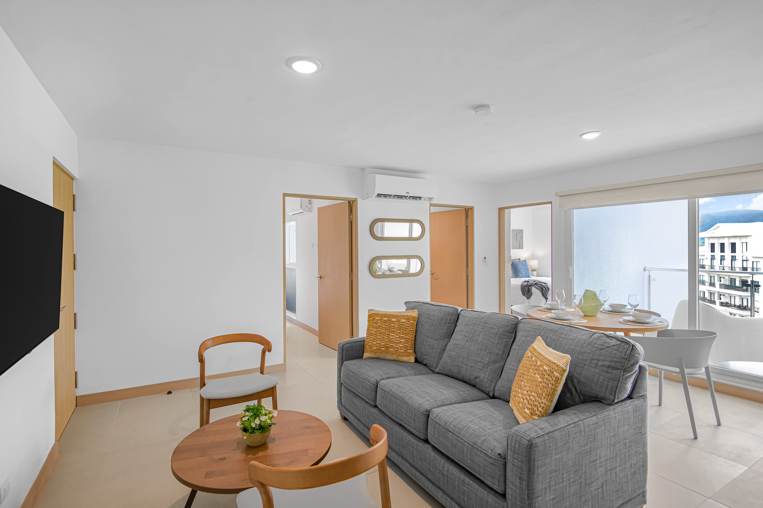 Bright living area featuring a gray sofa with yellow accent pillows, a round wooden coffee table, and a dining table visible through a doorway. An air conditioning unit is mounted above a decorative mirror on the wall.