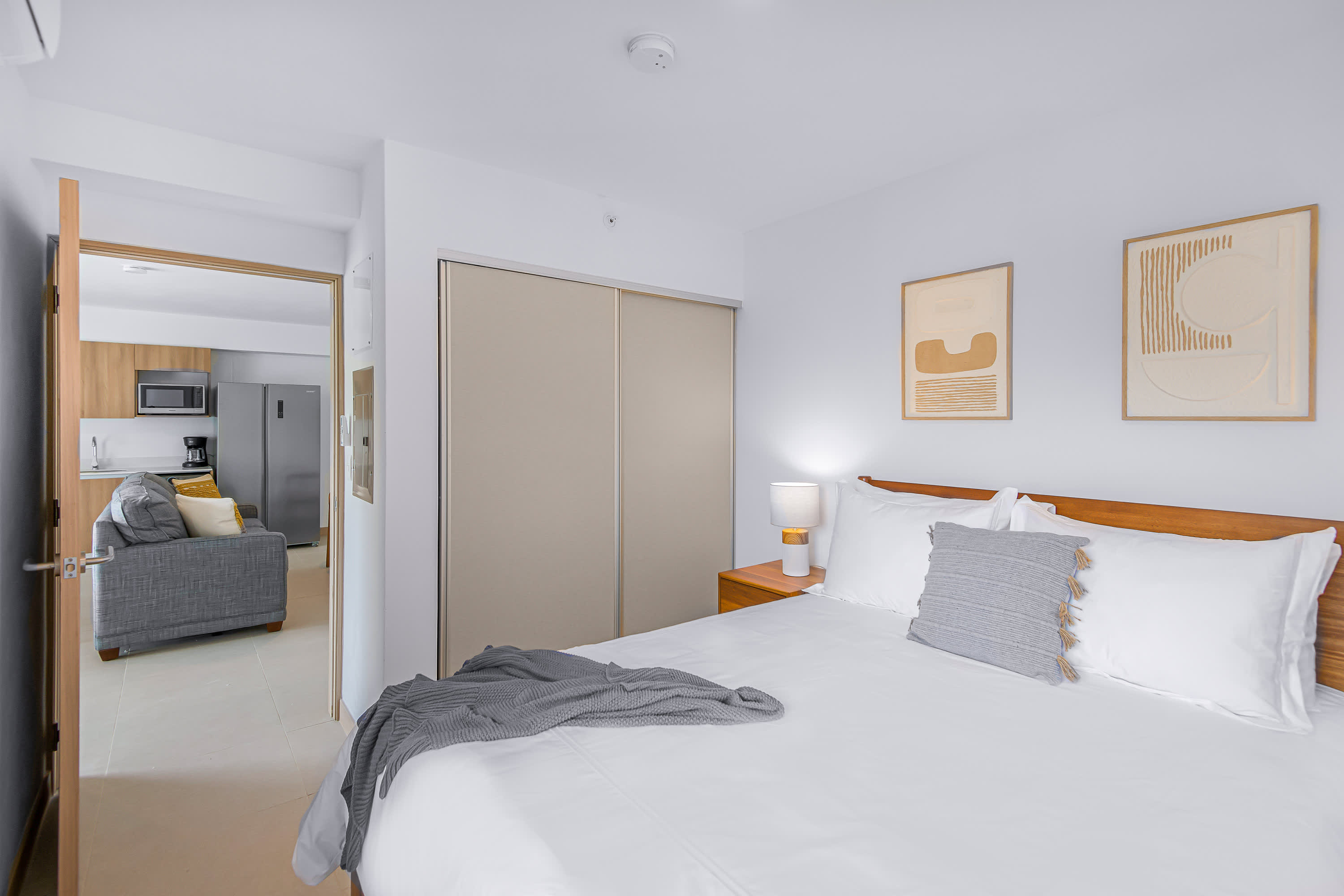 Bedroom with a bed, nightstands, and abstract art, showing an open doorway leading to the living room and kitchen area. A large sliding closet is visible on the left.