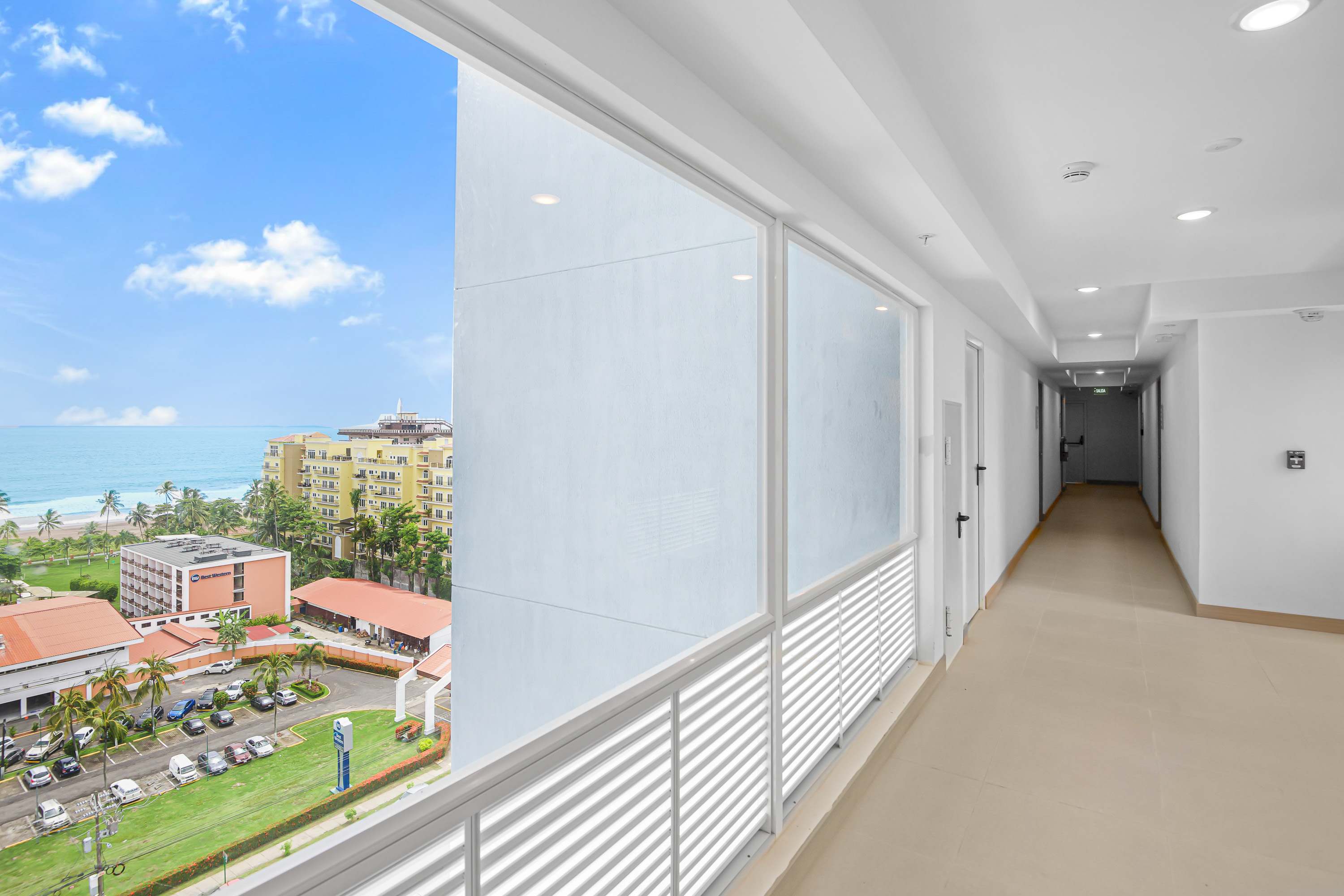 Bright interior hallway with light-colored tiled floors and white walls, featuring large windows that offer a partial view of the ocean and surrounding buildings.