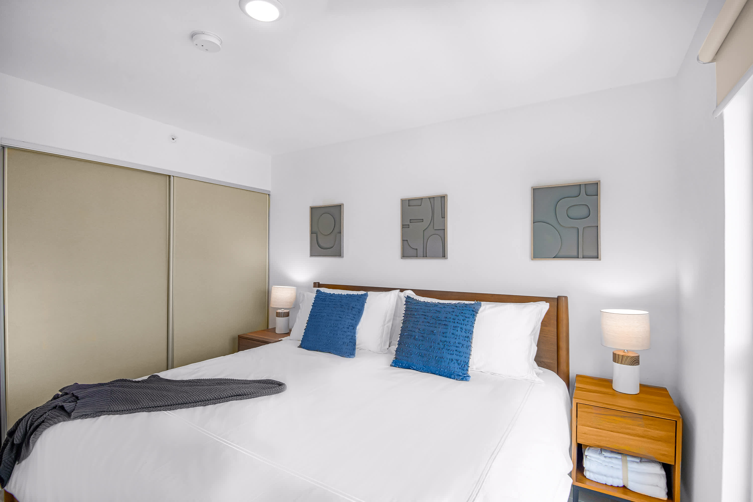 Spacious bedroom featuring a large bed with white linens and blue accent pillows, flanked by wooden nightstands with lamps. A large sliding closet door is visible, and abstract art hangs above the bed.