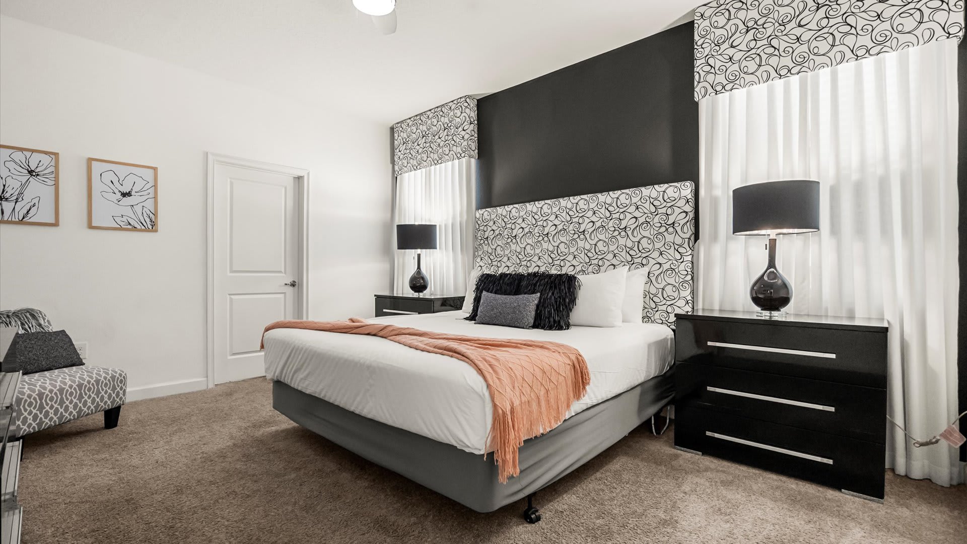 A stylish bedroom with a queen-sized bed featuring a patterned headboard and a black accent wall. Black nightstands with matching lamps flank the bed, and a peach throw blanket adds a pop of color.