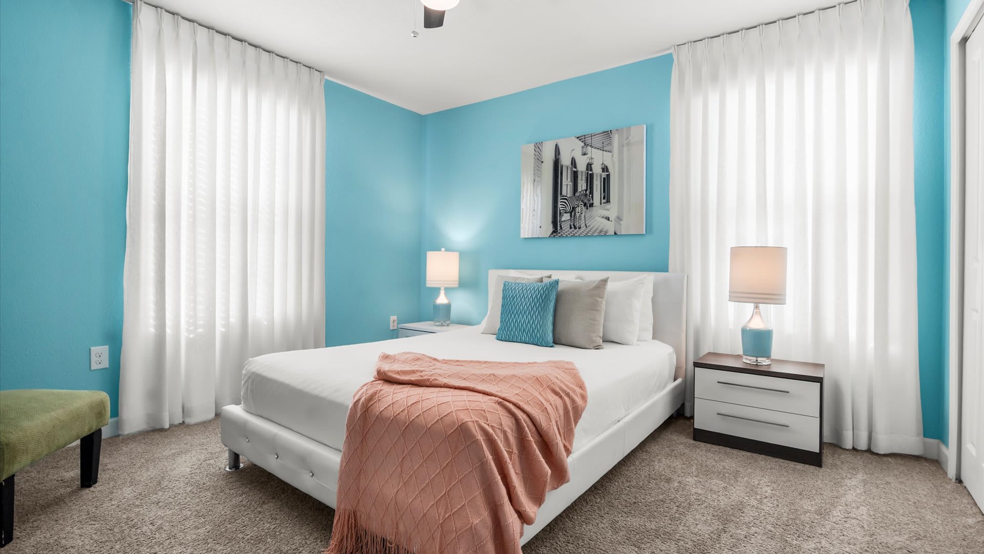 Vibrant bedroom with bright blue walls, a white upholstered bed, and sheer white curtains framing large windows. Two bedside tables with lamps flank the bed, and a large abstract artwork hangs above.