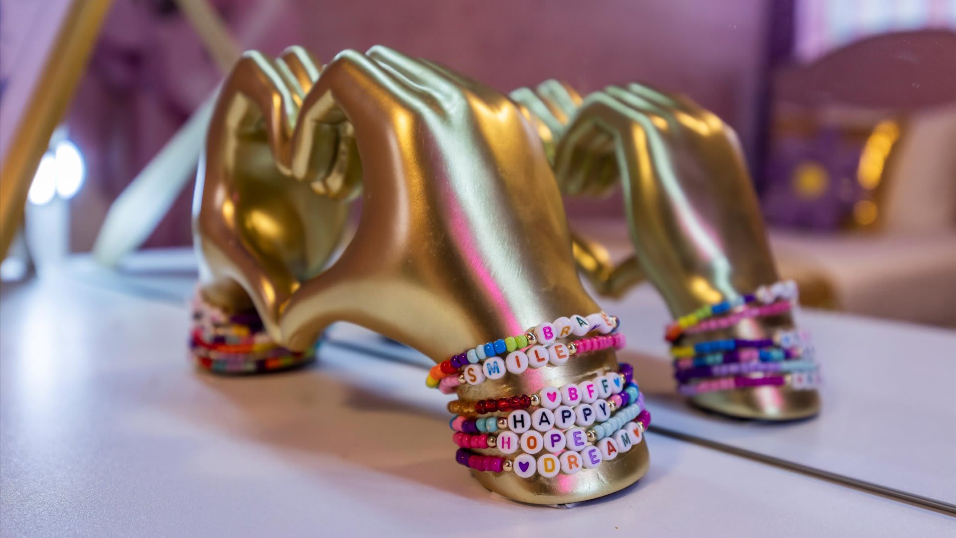 Close-up of colorful beaded bracelets with letter beads spelling words like 'SMILE' and 'HAPPY', displayed on golden hand sculptures on a white vanity surface.