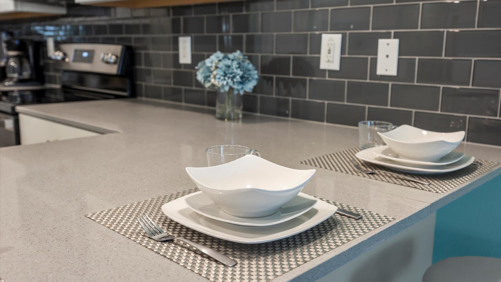 A close-up of the kitchen counter shows a light gray quartz countertop with two place settings, each featuring a square white plate, a bowl, and a glass, set on patterned placemats, with a dark gray subway tile backsplash in the background.