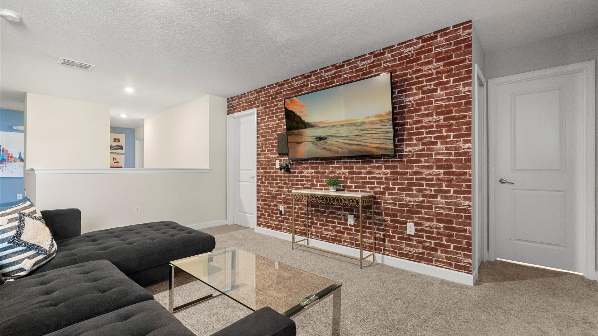 A wall-mounted flat-screen TV displays a beach scene above a gold console table, set against a red brick accent wall, with a dark gray sectional sofa and glass coffee table visible on the beige carpet.