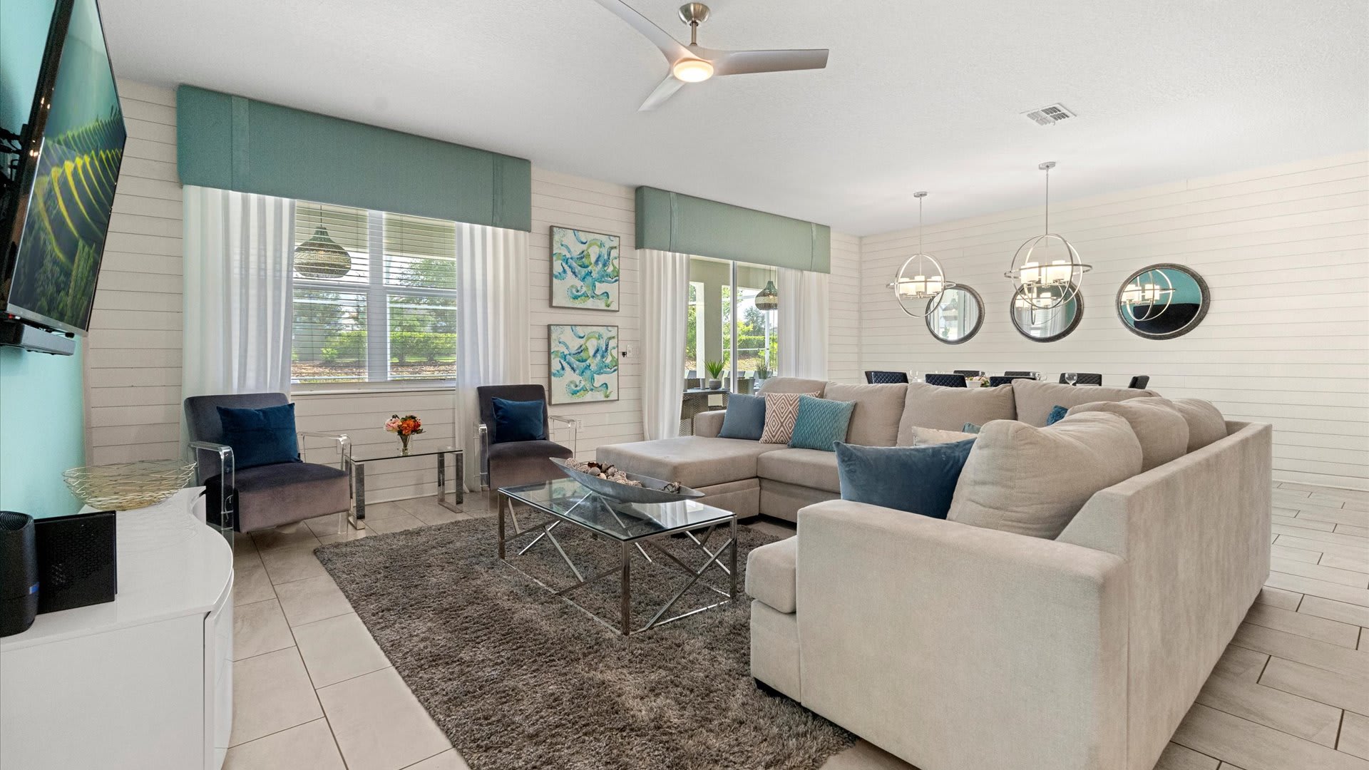 A spacious living room showcases a large light gray sectional sofa, a glass-top coffee table, and two clear acrylic accent chairs. White shiplap walls are complemented by teal window valances, and a ceiling fan provides comfort.