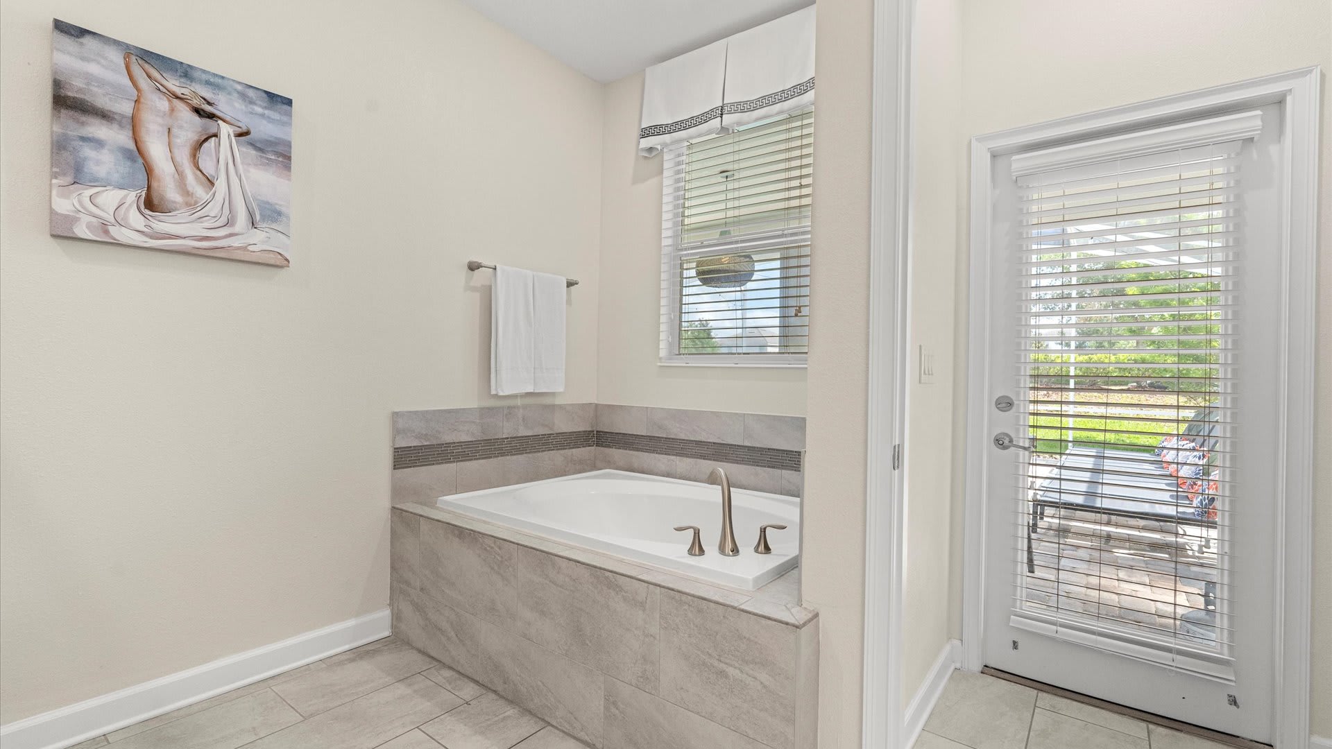 A spacious bathroom area featuring a built-in bathtub with gray tiled surround and a window with blinds above it. A door with blinds provides access to an outdoor space, complementing the light-colored walls.