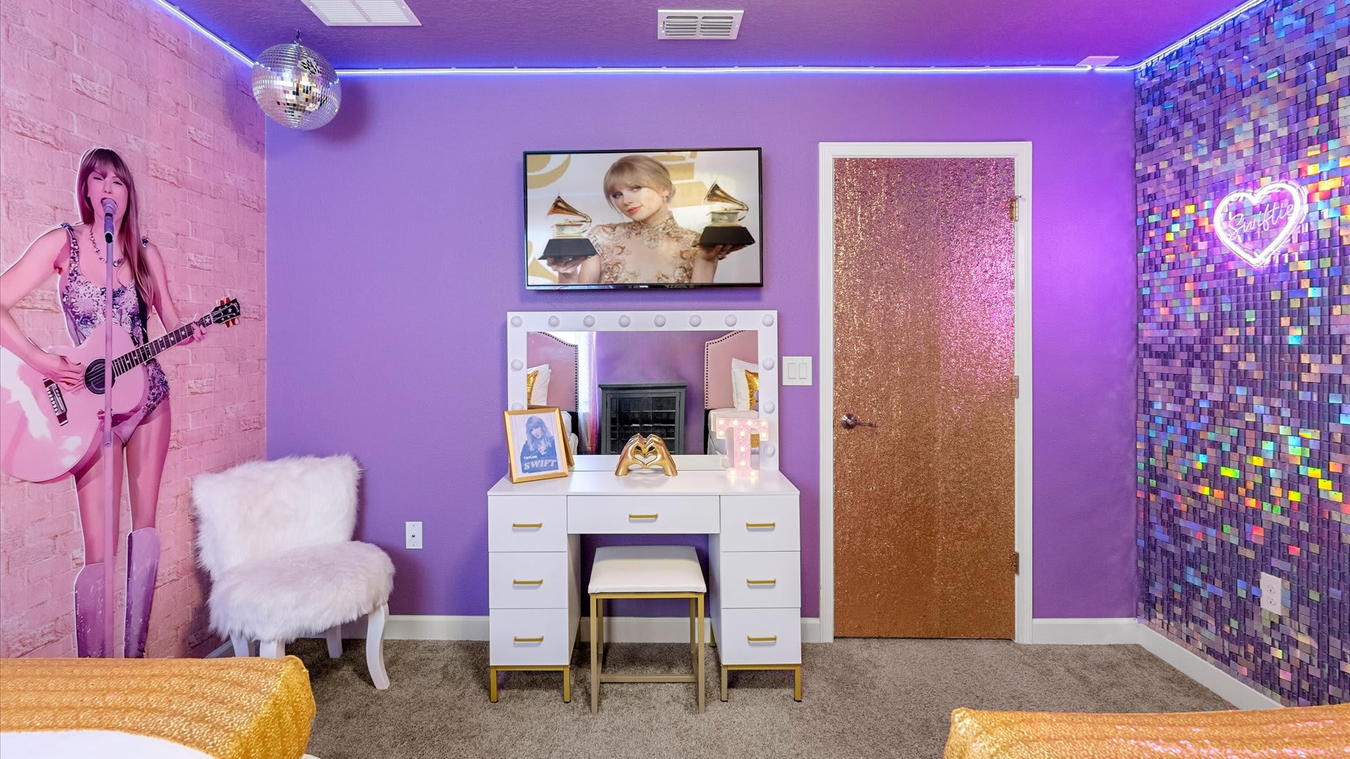 Another angle of the Taylor Swift themed bedroom, featuring a white vanity with a lighted mirror, a wall-mounted TV, and a large Taylor Swift cutout. A shimmering gold door adds to the room's unique style.