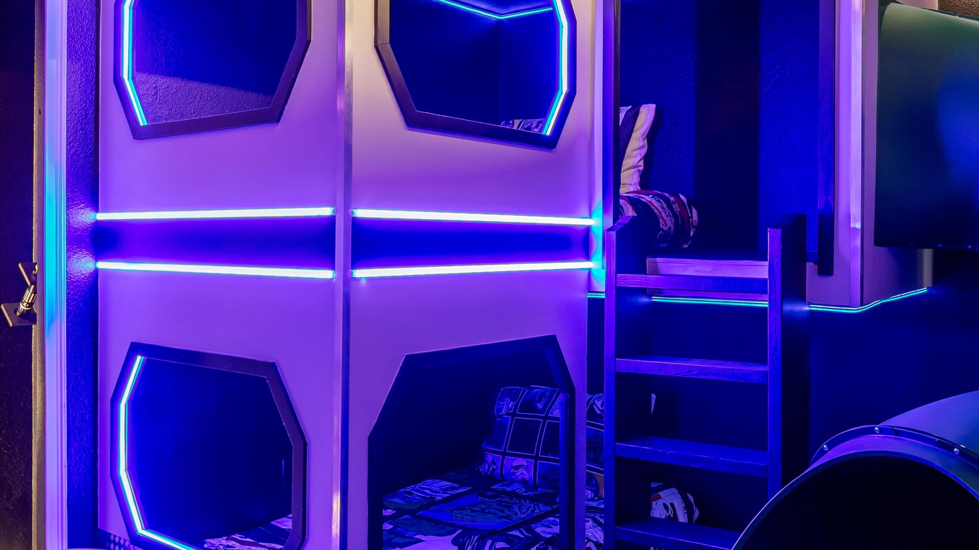 A themed bedroom featuring futuristic bunk beds designed as sleeping pods, illuminated by striking blue and purple LED strip lighting, with a wooden ladder providing access to the upper bunk.