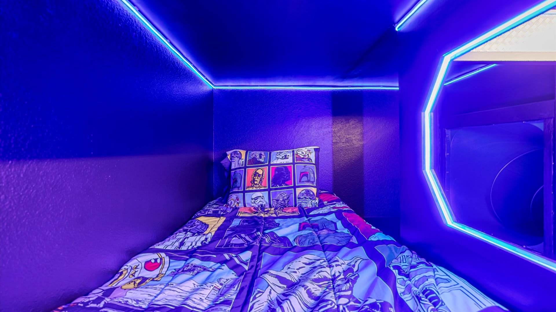 A cozy, themed sleeping pod within a bunk bed structure, featuring a Star Wars-themed comforter and pillow, brightly outlined by blue LED strip lighting.