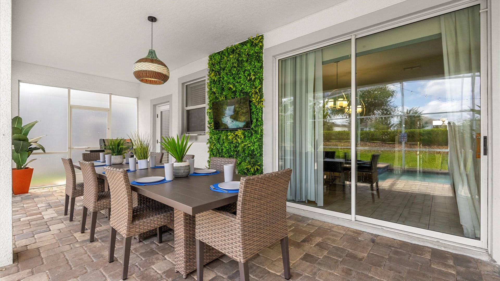 An outdoor patio features a long rectangular dining table with wicker chairs, set on a brick paver floor, with a green wall accent and a view of a screened-in pool area through sliding glass doors.