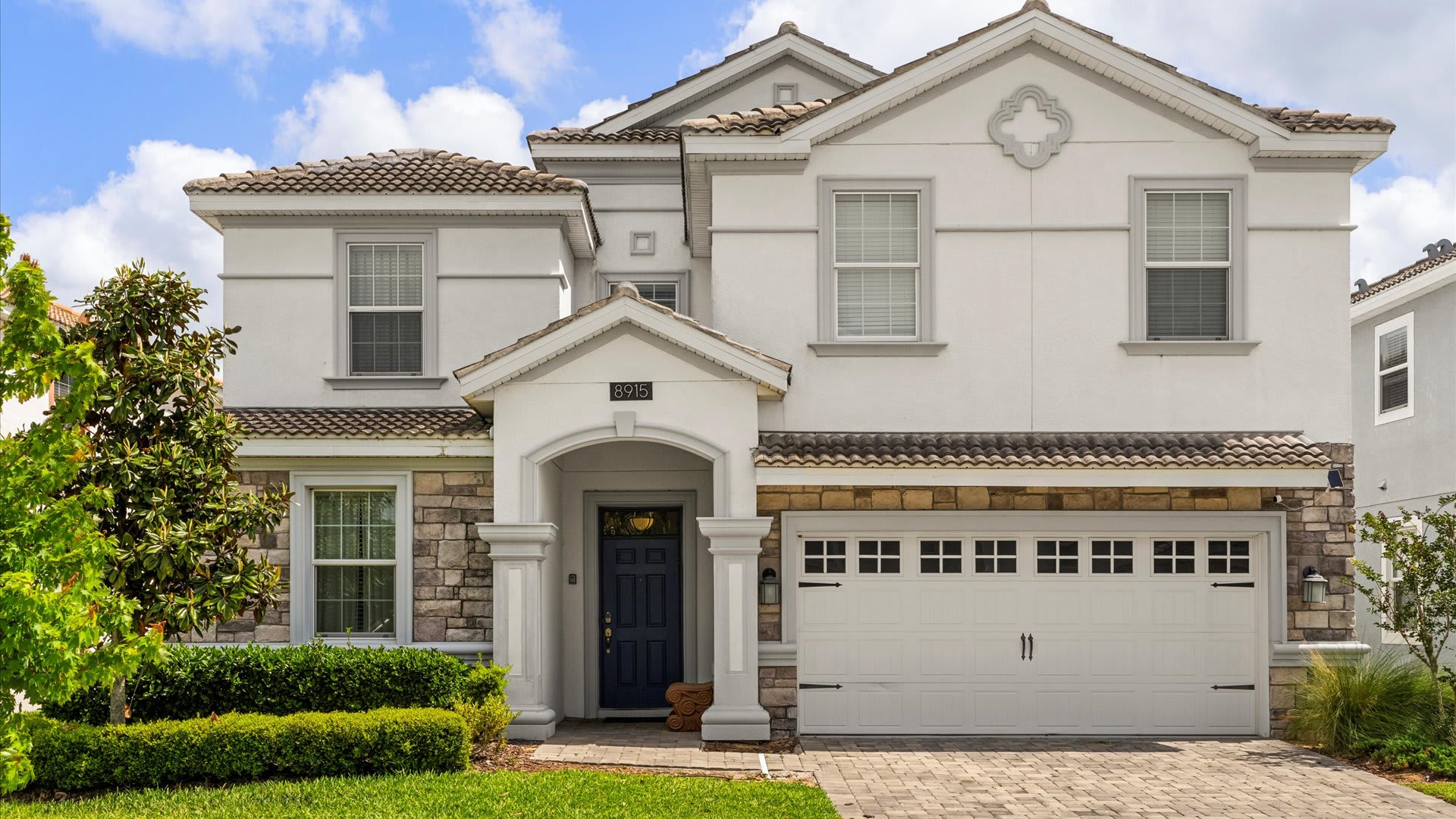 A modern two-story house with a light grey stucco exterior and stone accents features a dark blue front door and a two-car garage. The front yard is neatly landscaped with green bushes and a paved driveway.