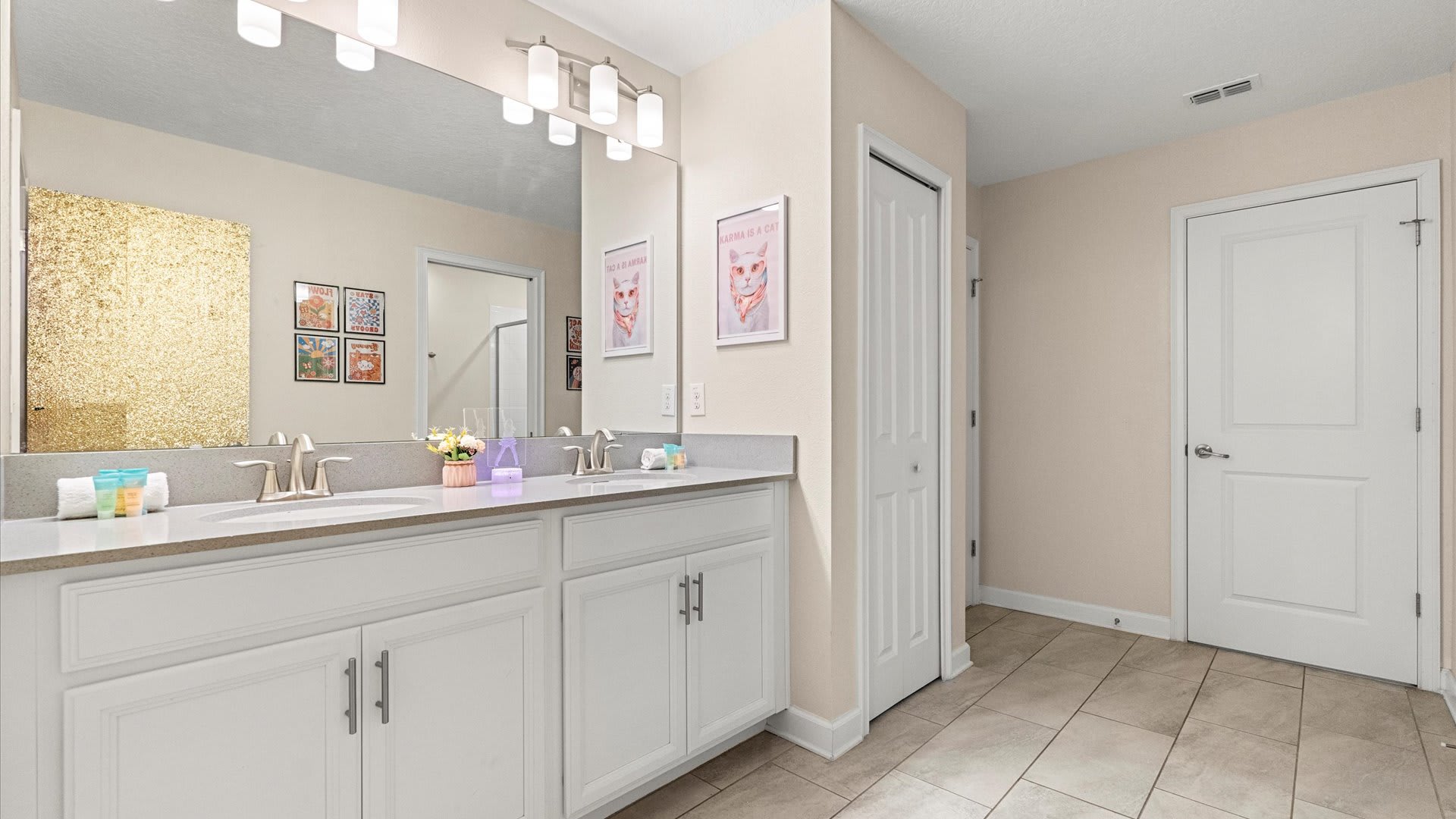 A bright bathroom featuring a double vanity with white cabinets and light gray countertops, a large mirror reflecting a gold sequin wall, and two framed "Karma is a Cat" posters on the wall.
