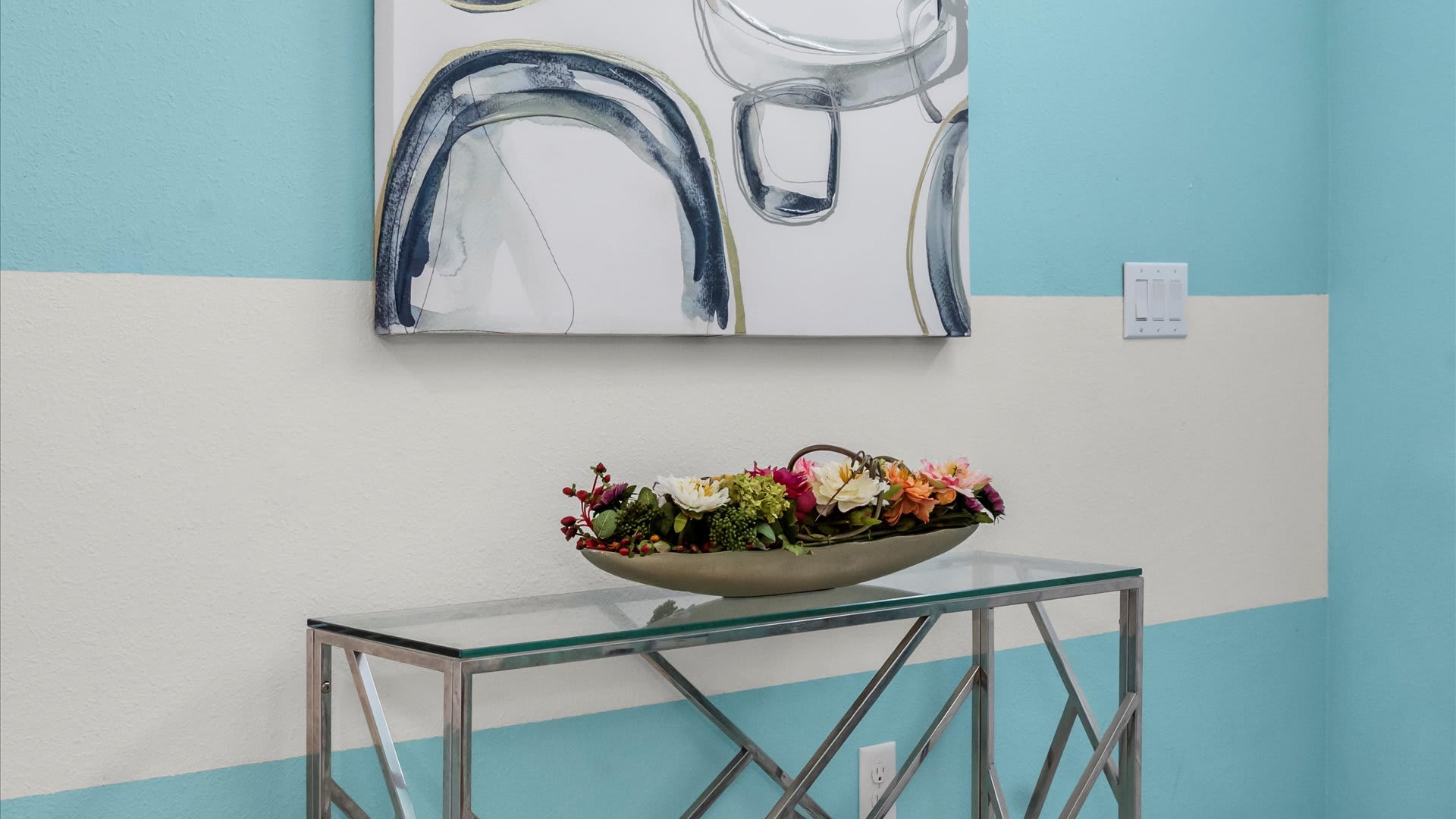 A console table with a glass top and metal base, adorned with a decorative bowl of artificial flowers, set against a wall with light blue and white horizontal stripes and an abstract painting above.