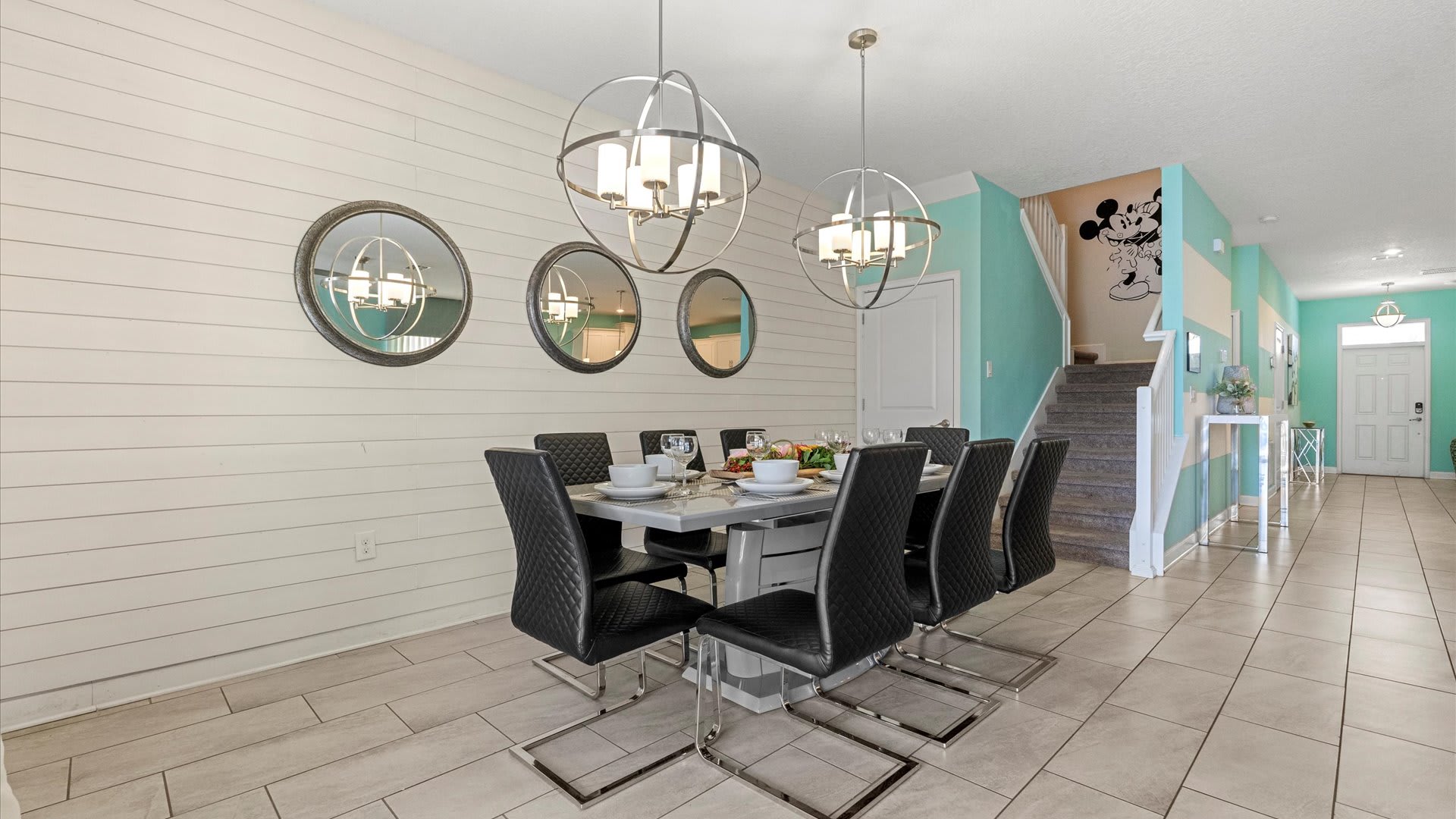 A contemporary dining room features a long gray table with eight black quilted chairs, illuminated by two spherical chandeliers. White shiplap walls are adorned with three round mirrors, and a staircase with a Mickey Mouse decal is visible in the background.