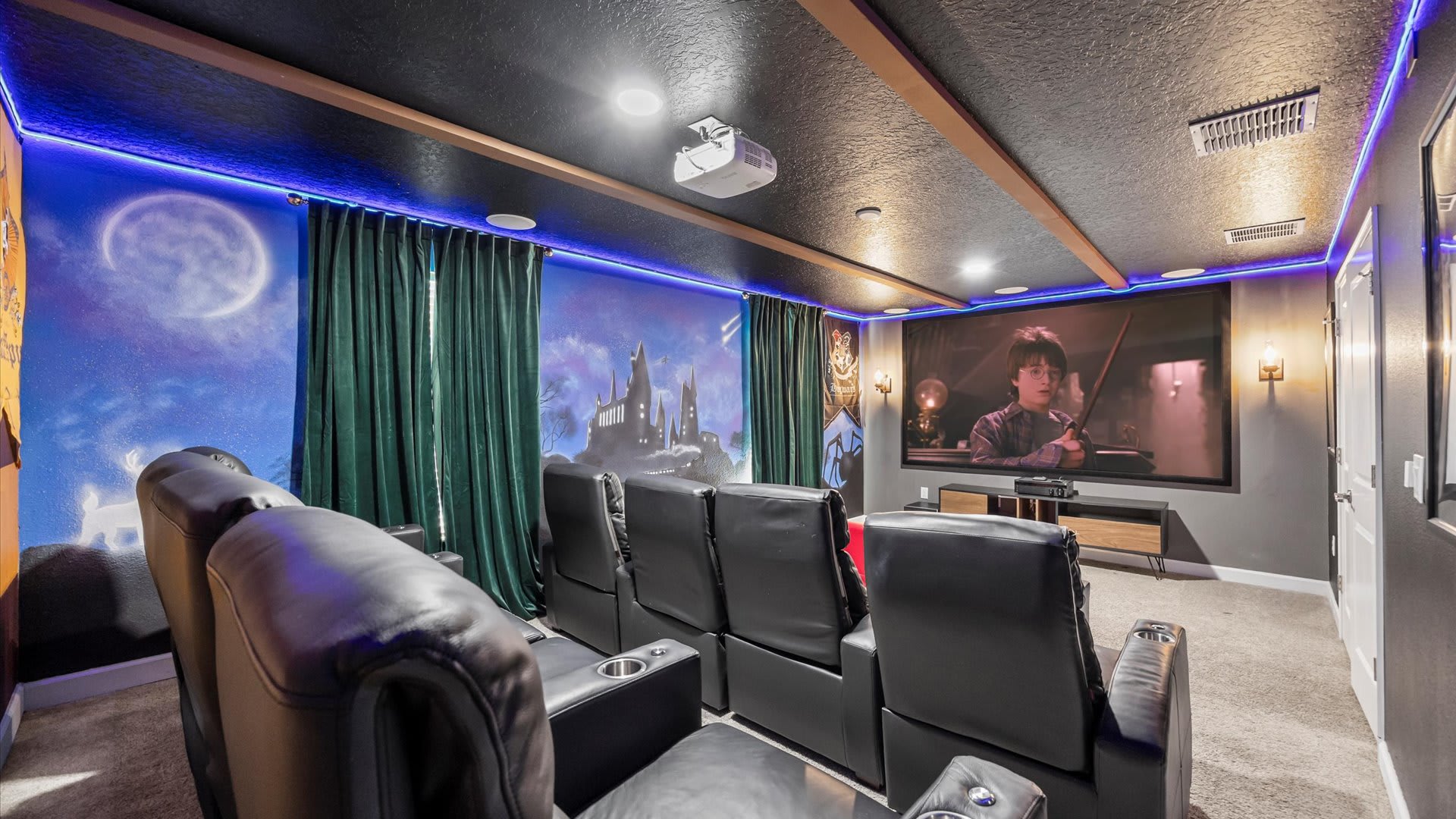 A Harry Potter-themed home cinema features comfortable black reclining seats, a large projection screen displaying a movie, and a mural of Hogwarts castle on the wall, illuminated by blue LED strip lighting.