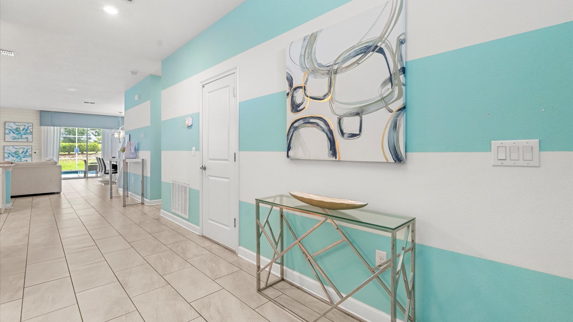 A bright and inviting hallway features eye-catching white and aqua striped walls, leading to various rooms. A modern glass-top console table with a metallic base holds a decorative bowl, and abstract artwork hangs above it.