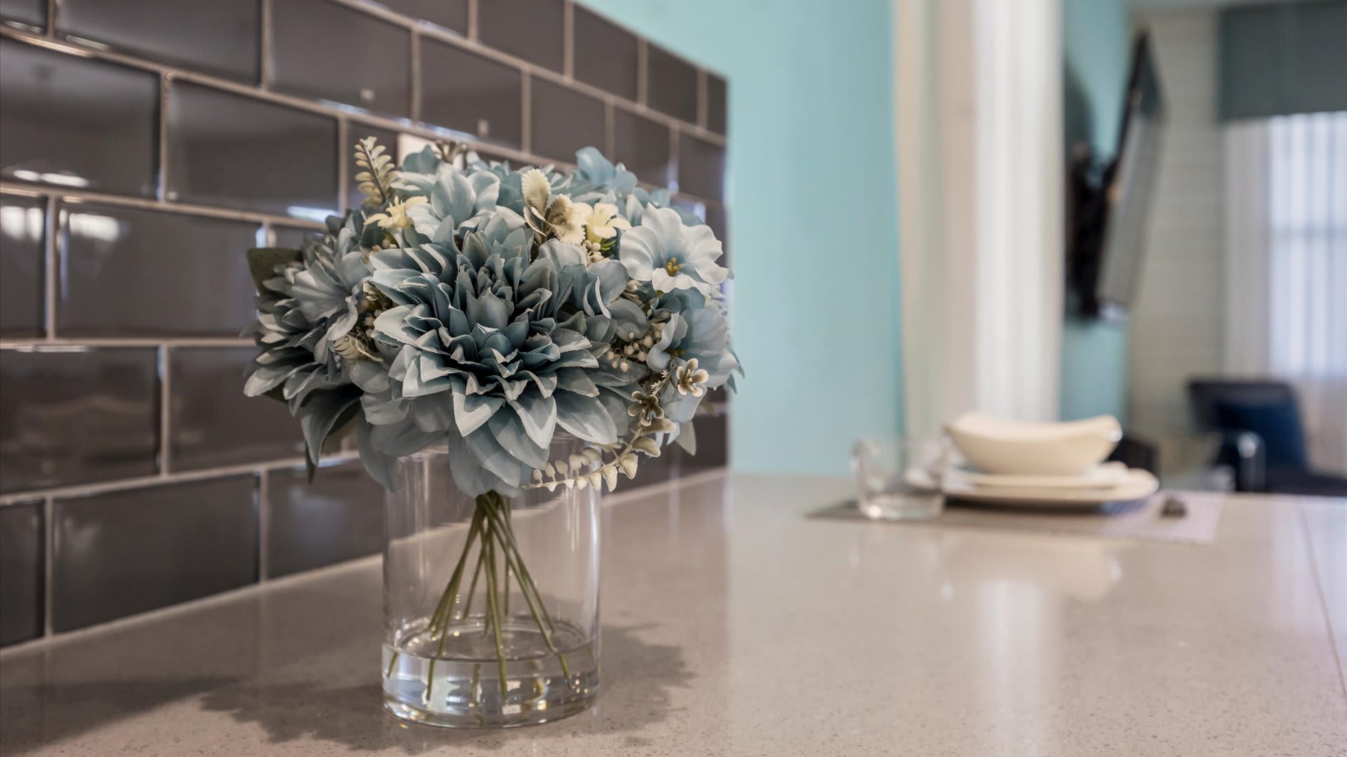 A close-up view of the kitchen countertop highlights a clear glass vase filled with light blue and white artificial flowers, set against a dark gray subway tile backsplash, with a light gray quartz countertop.