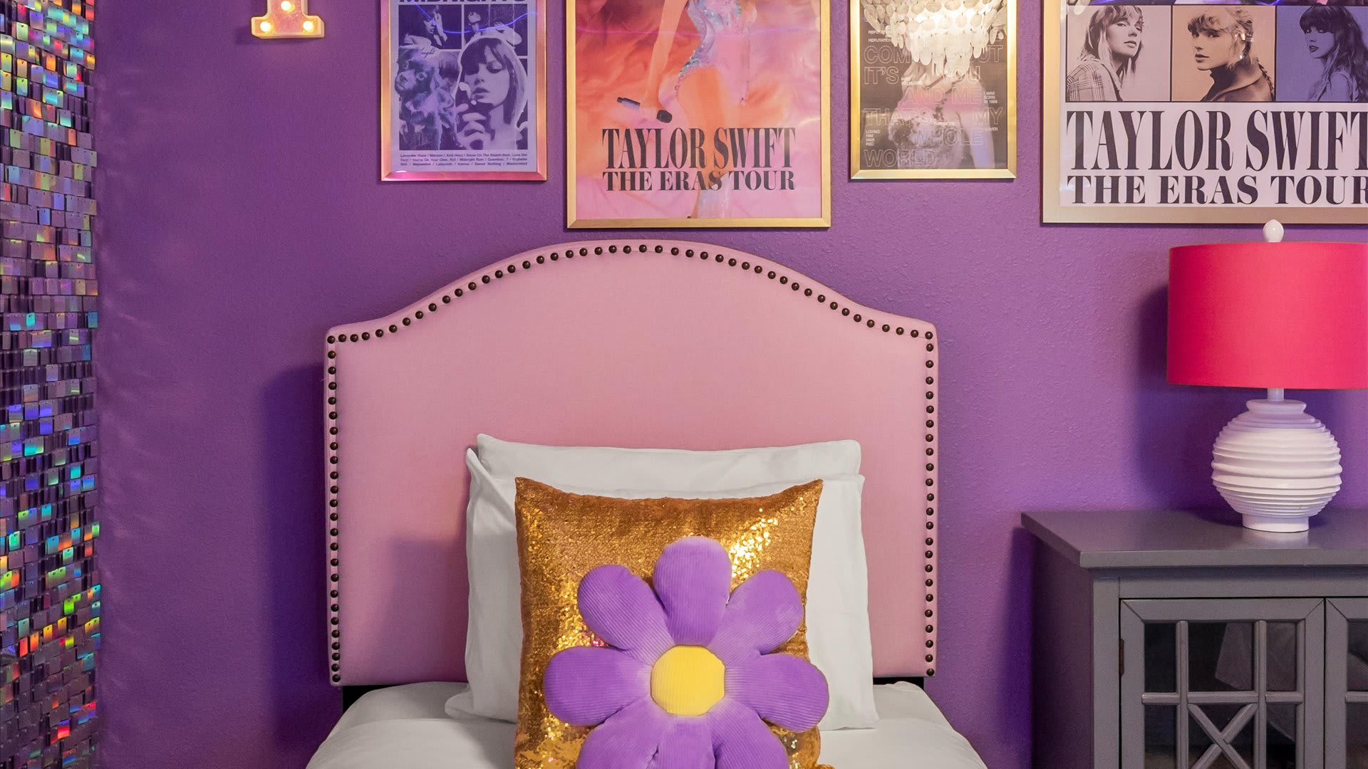 Close-up of one of the pink upholstered twin beds in the Taylor Swift themed room, featuring a gold sequin pillow with a purple flower and a bright pink lamp on a grey nightstand.