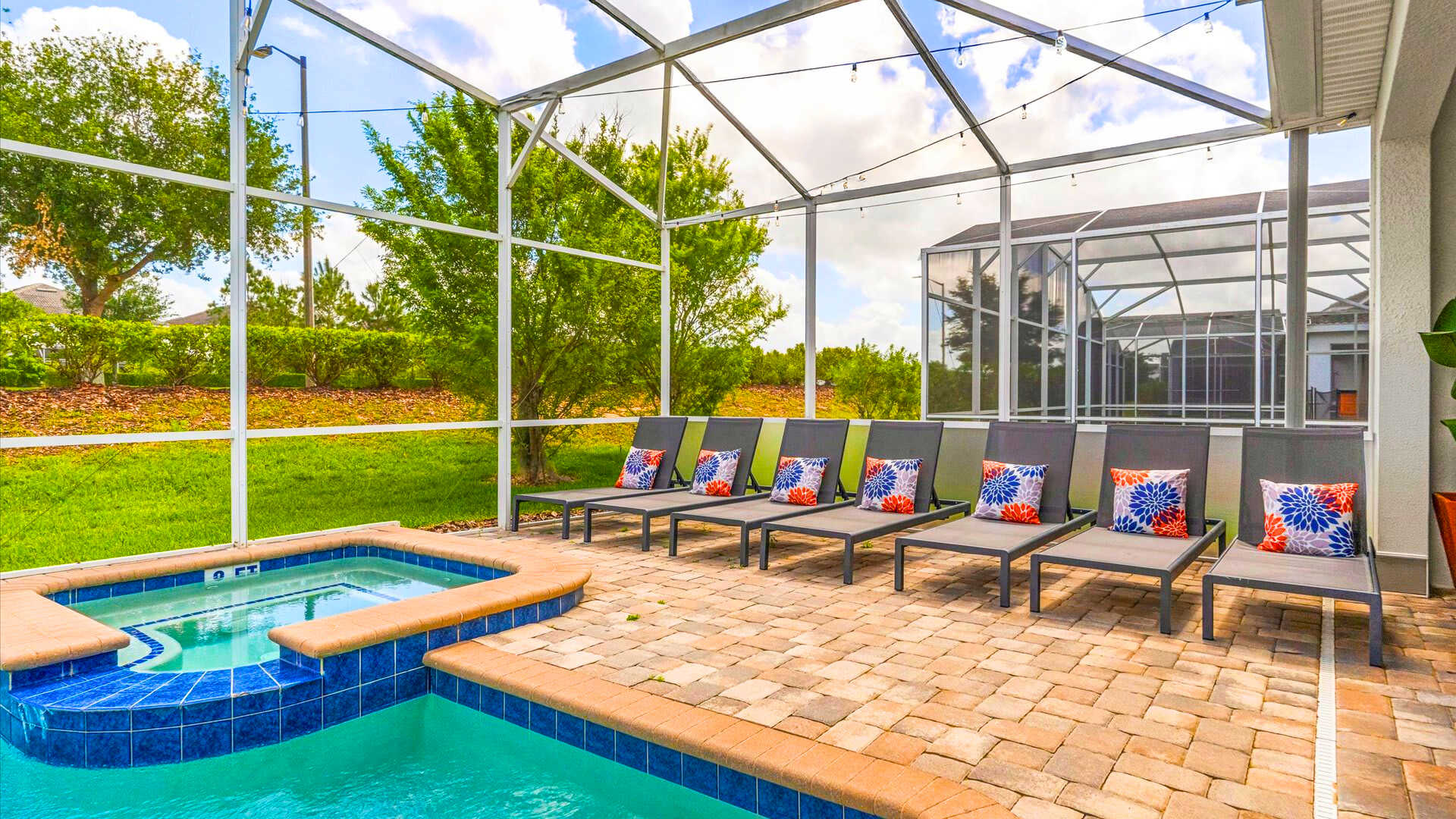 A close-up view of the private pool and hot tub area, surrounded by brick pavers and several dark lounge chairs with bright floral pillows. The screened enclosure allows for open views of the grassy backyard.