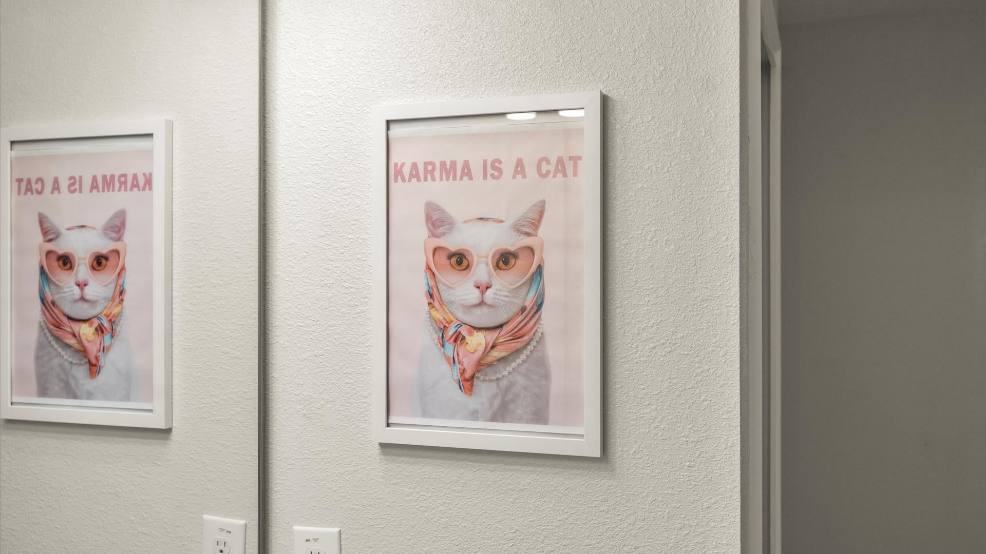 A close-up of a framed poster on a white wall, depicting a white cat wearing pink heart-shaped sunglasses and a colorful scarf, with the text "KARMA IS A CAT" above it.