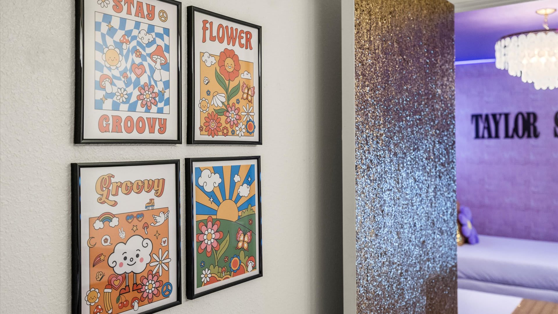 A white wall adorned with four framed, colorful groovy-themed posters, adjacent to a shimmering gold sequin wall that leads into a purple-lit room with "TAYLOR S" visible, indicating a themed bedroom.