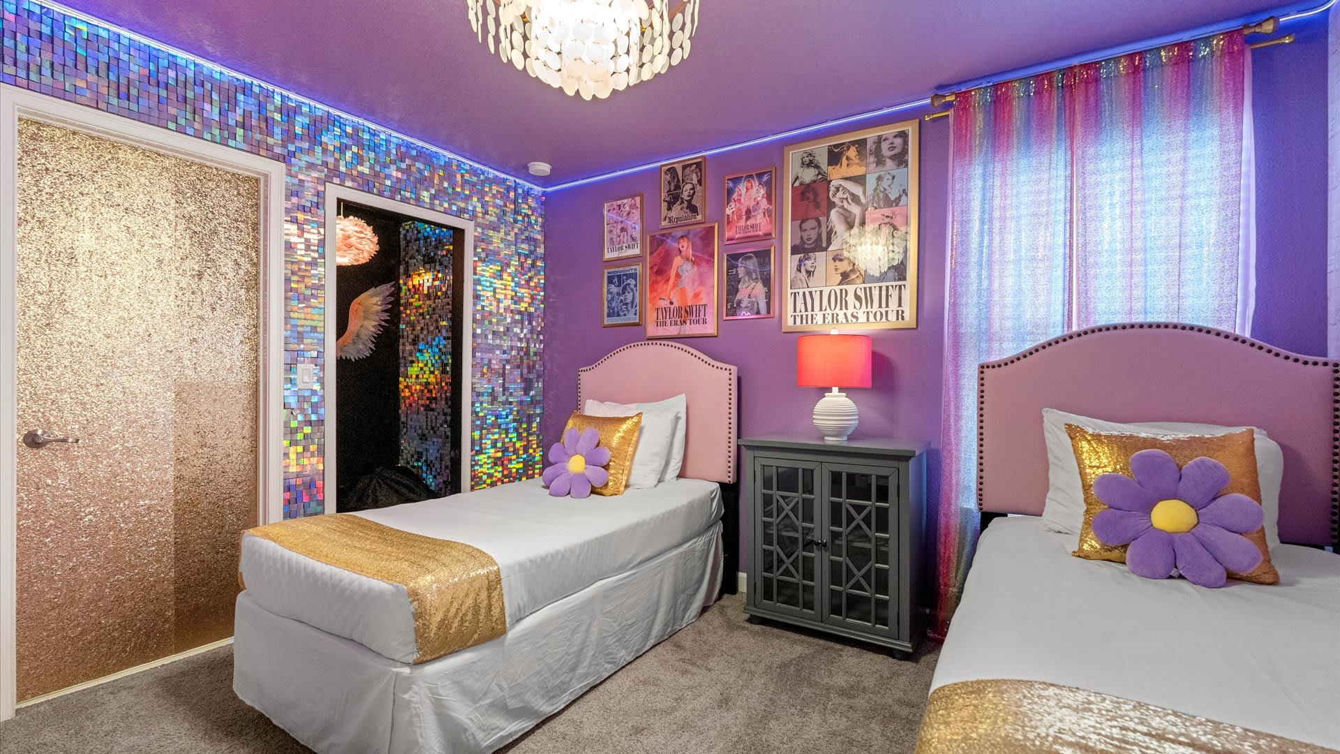 View of the Taylor Swift themed bedroom showcasing a shimmering, iridescent mirrored wall and a matching closet door. Two pink twin beds with gold accents are visible, along with wall posters and a chandelier.