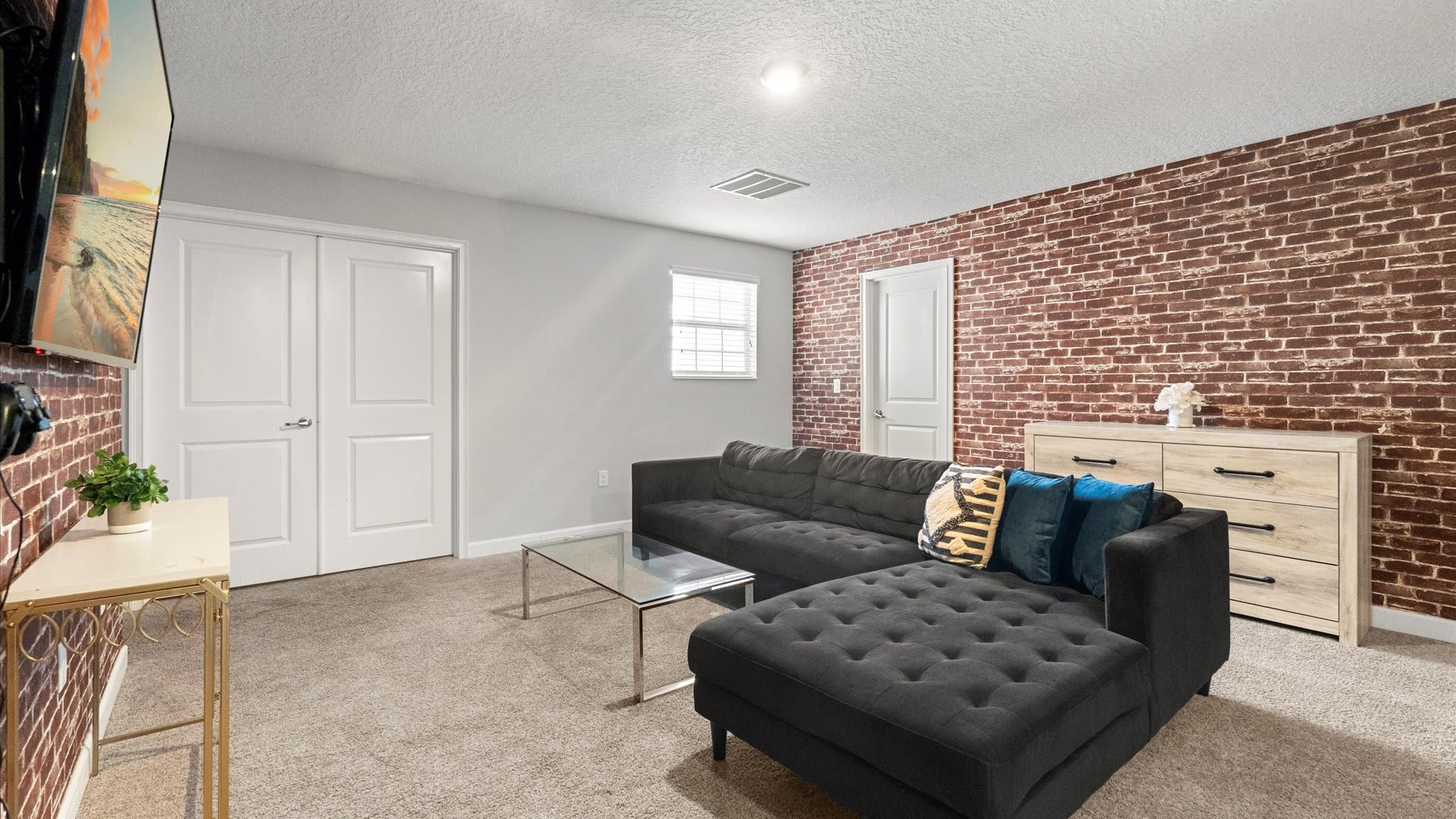 A spacious media room showcases a dark gray sectional sofa with patterned and blue throw pillows, a glass coffee table, and a light wood dresser, all on a beige carpet, with a red brick accent wall and white doors.