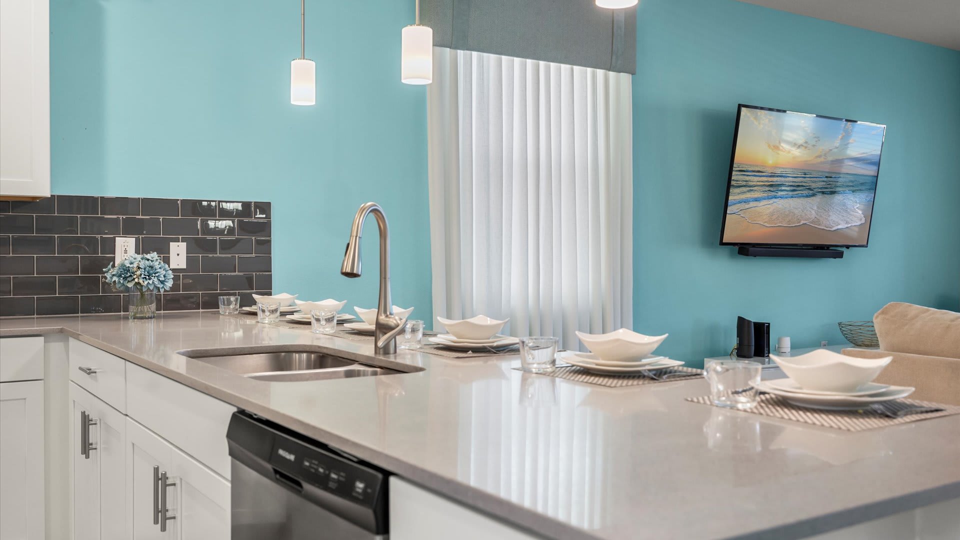 A close-up of a kitchen island features a sleek stainless steel sink and faucet, neatly set with white dinnerware and striped placemats. A wall-mounted TV is visible in the background, complementing the light gray countertops and aqua walls.