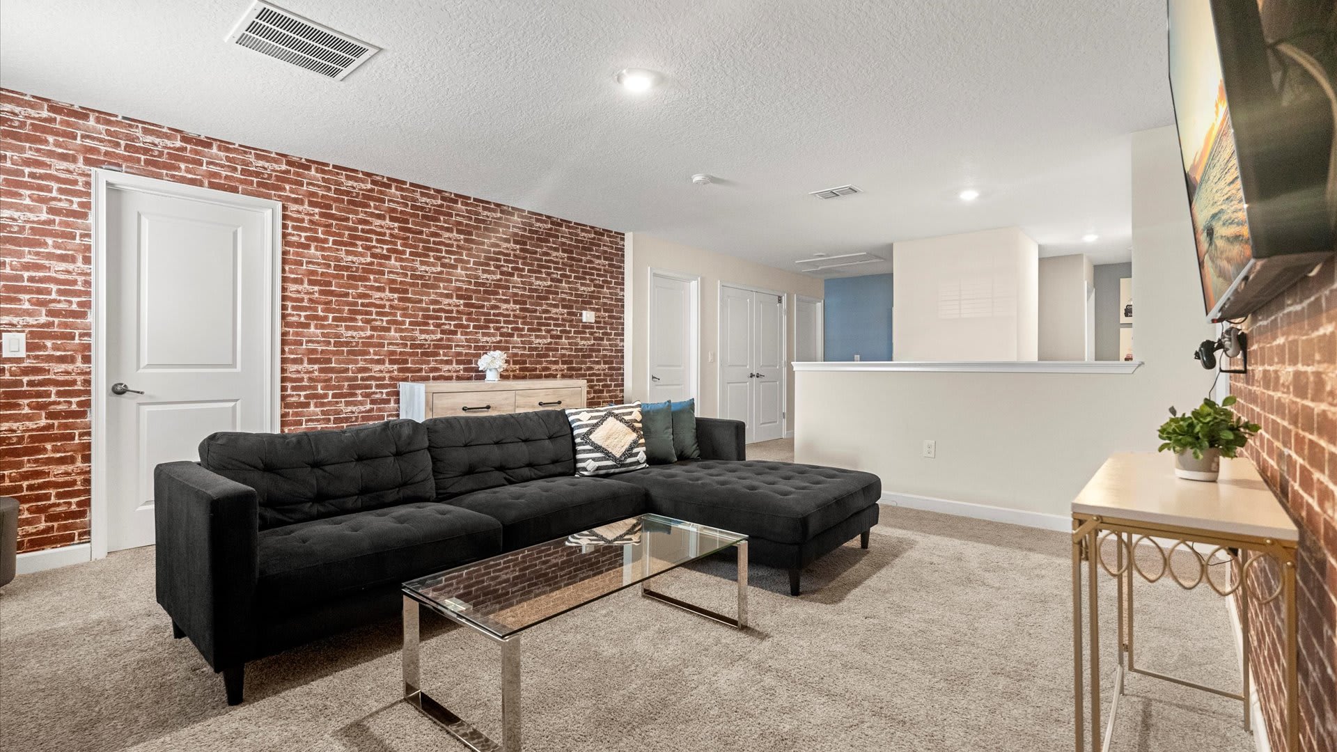 A cozy media room features a large dark gray sectional sofa with throw pillows, a glass coffee table, and a light wood dresser, all on a beige carpet, with a striking red brick accent wall and white doors.