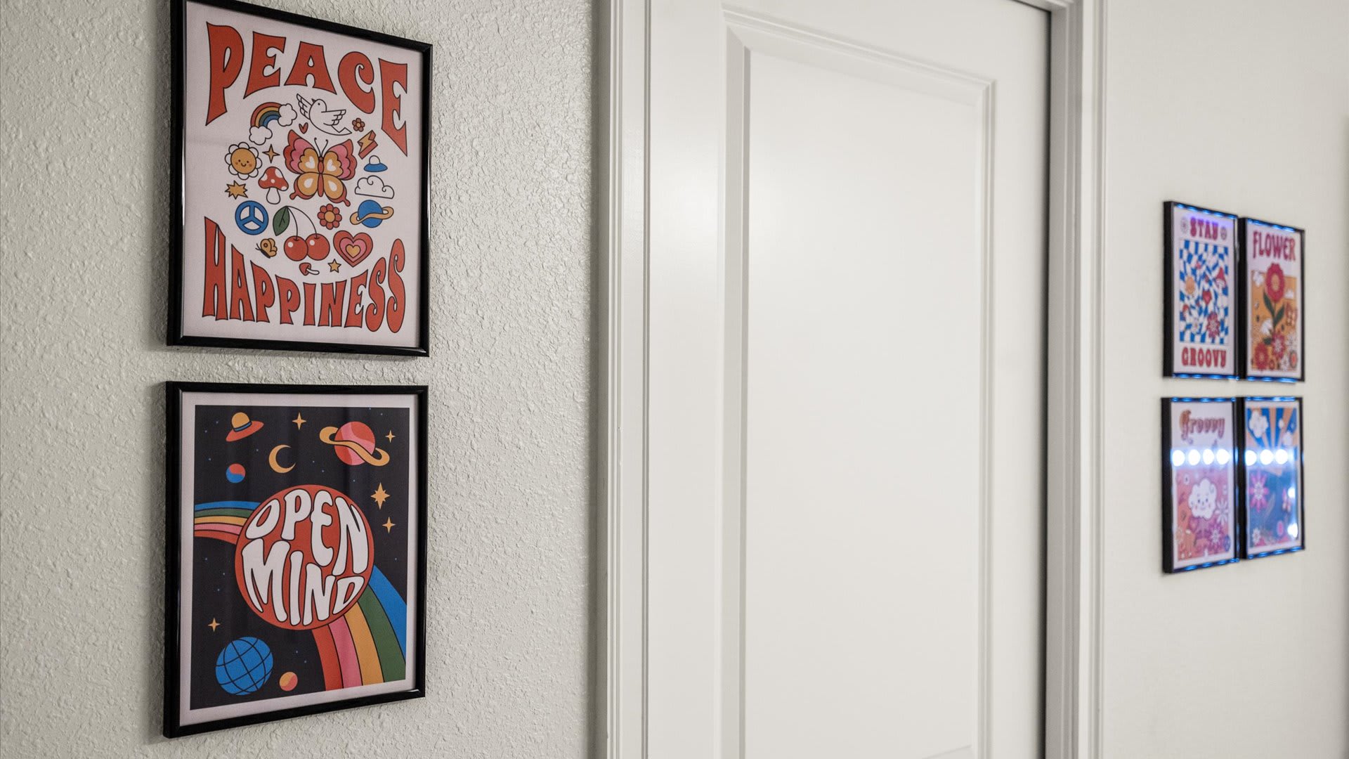 A white wall displaying two framed, colorful posters with retro-inspired designs, one reading "PEACE HAPPINESS" with a butterfly, and another "OPEN MIND" with a rainbow and planets.