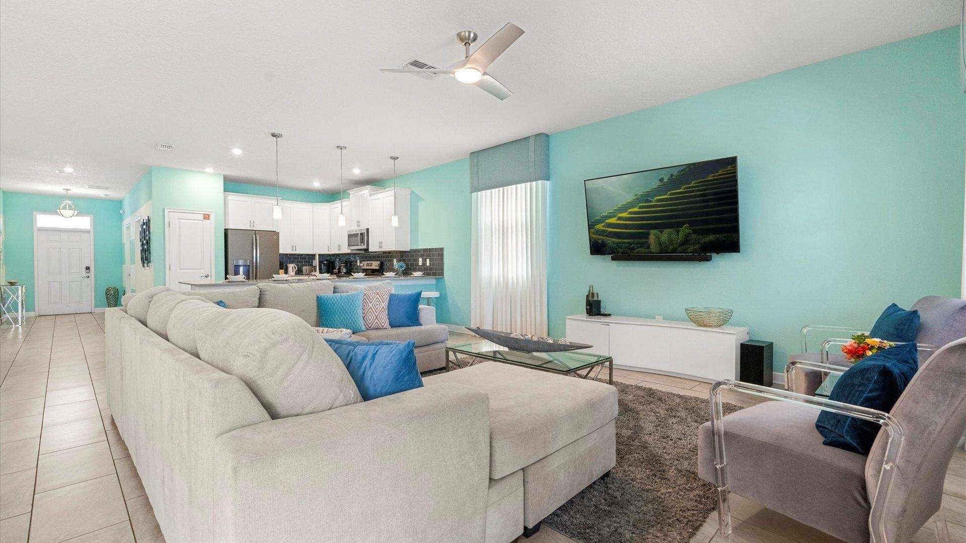 A bright living room features a large L-shaped light gray sectional sofa, a glass coffee table, and a wall-mounted TV displaying a landscape. Vibrant aqua blue and white walls create a lively atmosphere, with a view into the modern kitchen.