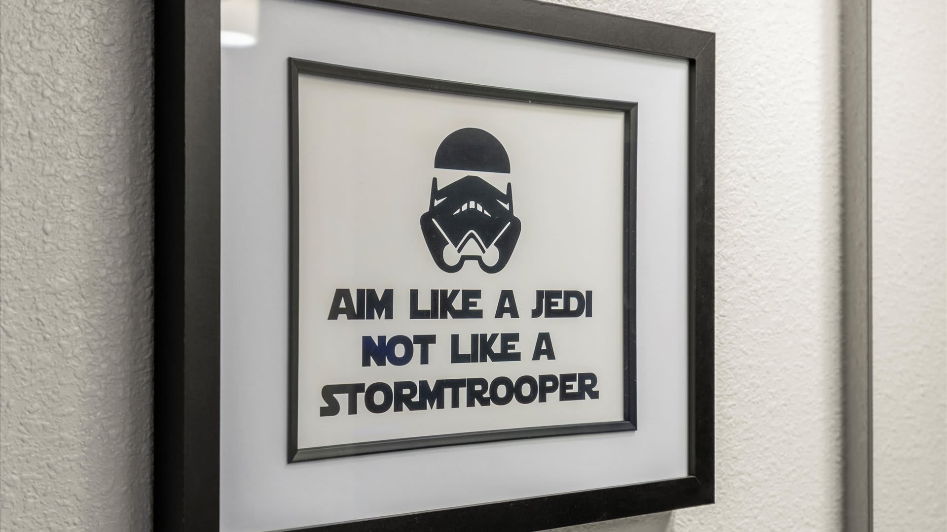 A close-up of a framed motivational sign in a bathroom, humorously stating 'AIM LIKE A JEDI NOT LIKE A STORMTROOPER' with a Stormtrooper helmet icon.