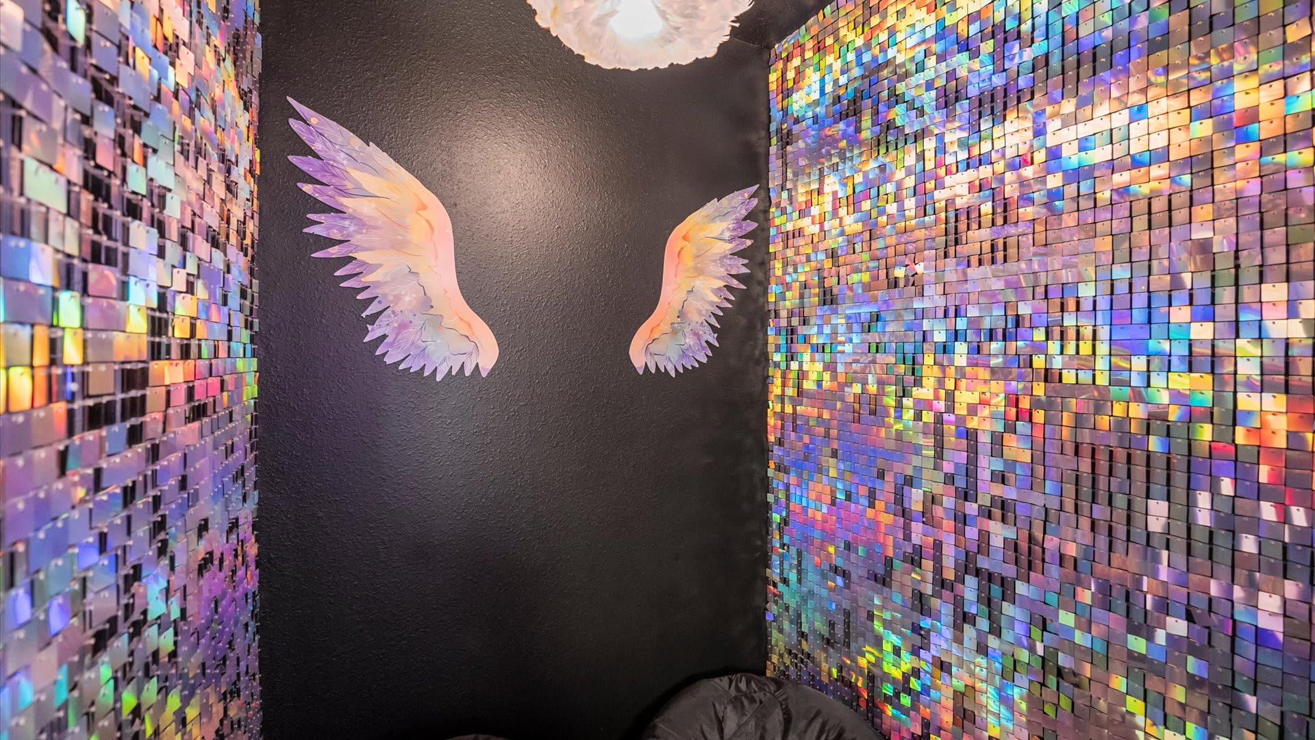 A vibrant themed room featuring iridescent sequin walls on two sides and a dark wall with large, colorful angel wings, creating a whimsical photo opportunity space.