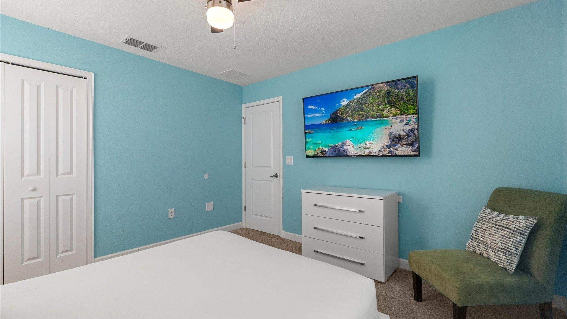 Another view of the vibrant blue bedroom, featuring a wall-mounted flat-screen TV displaying a beach scene, a white dresser with silver pulls, and a green accent chair in the corner.