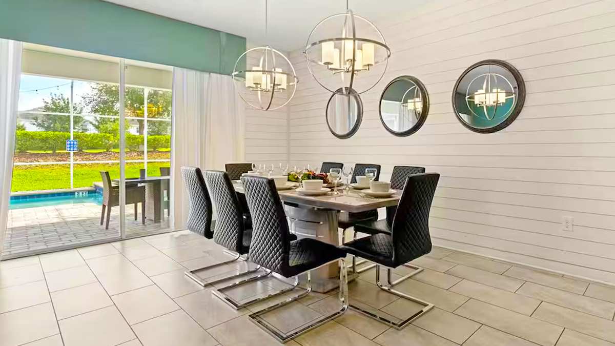 A modern dining area features a long gray table set for eight, with stylish black quilted chairs on chrome bases. Two large spherical chandeliers illuminate the space, and a sliding glass door opens to a patio with a pool view.