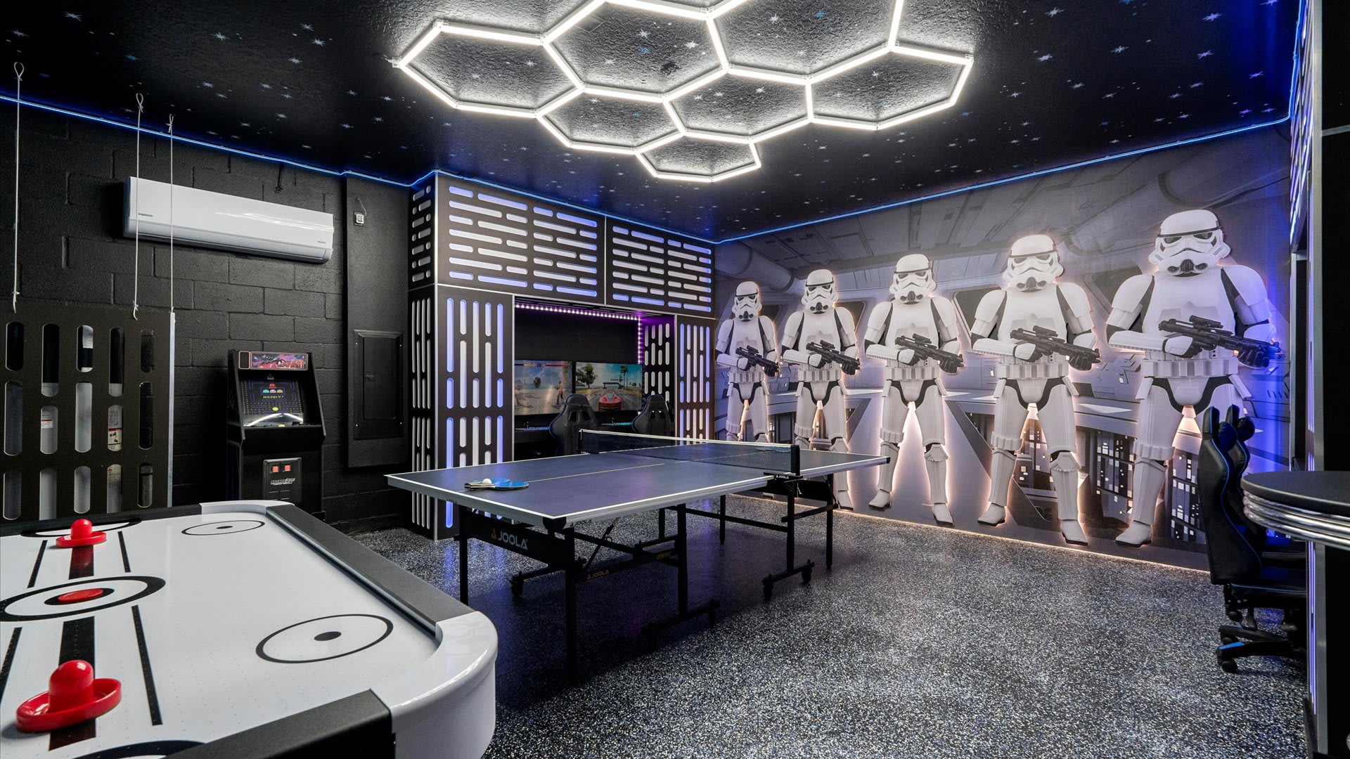A spacious Star Wars-themed game room features a ping-pong table, an air hockey table, and an arcade machine, with life-sized Stormtrooper figures lining a mural and hexagonal LED ceiling lights illuminating the space.