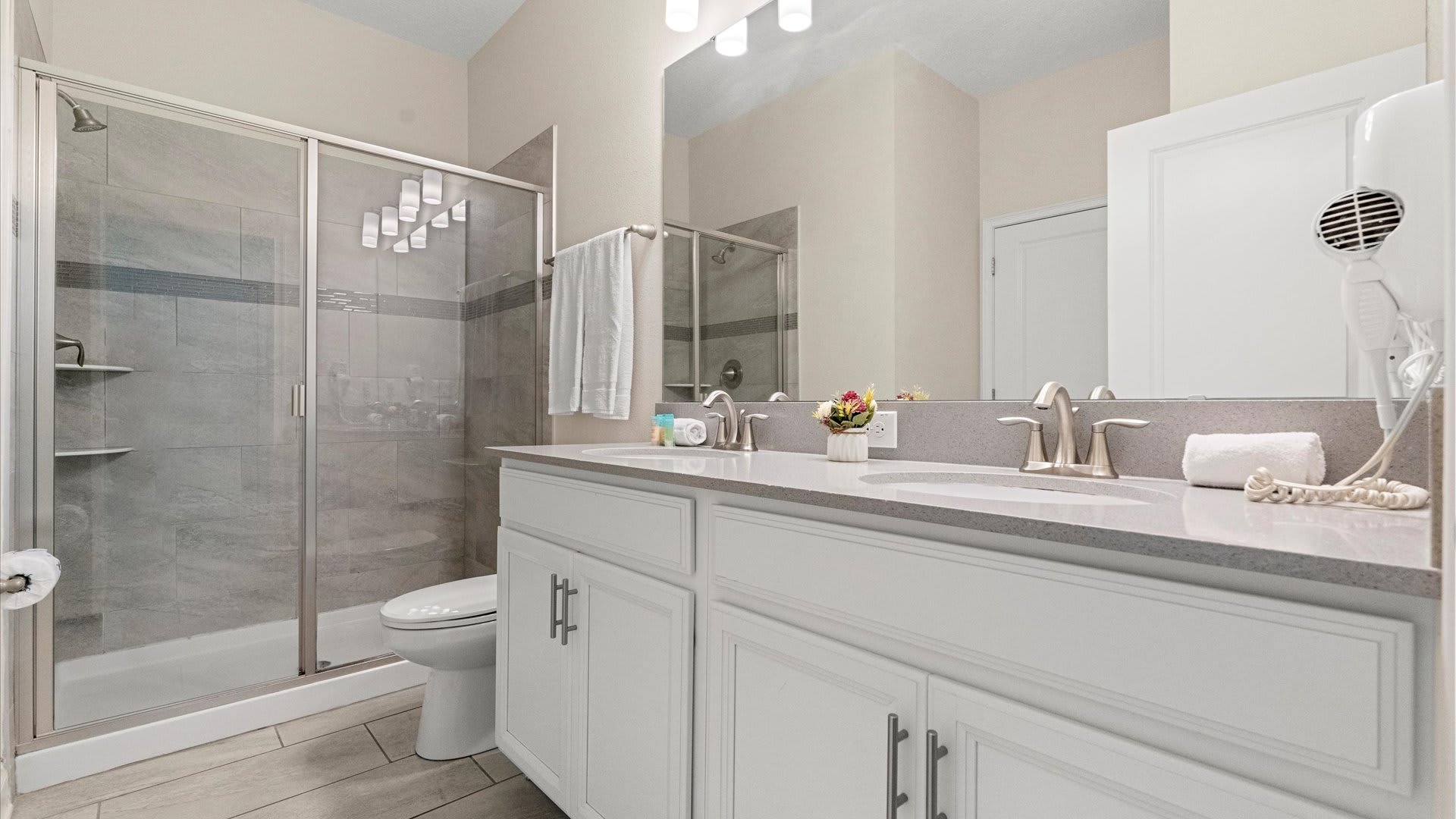 A modern bathroom featuring a double vanity with a light gray countertop and white cabinets, alongside a glass-enclosed walk-in shower with gray tiled walls. A large mirror extends above the vanity, reflecting the bright lighting.