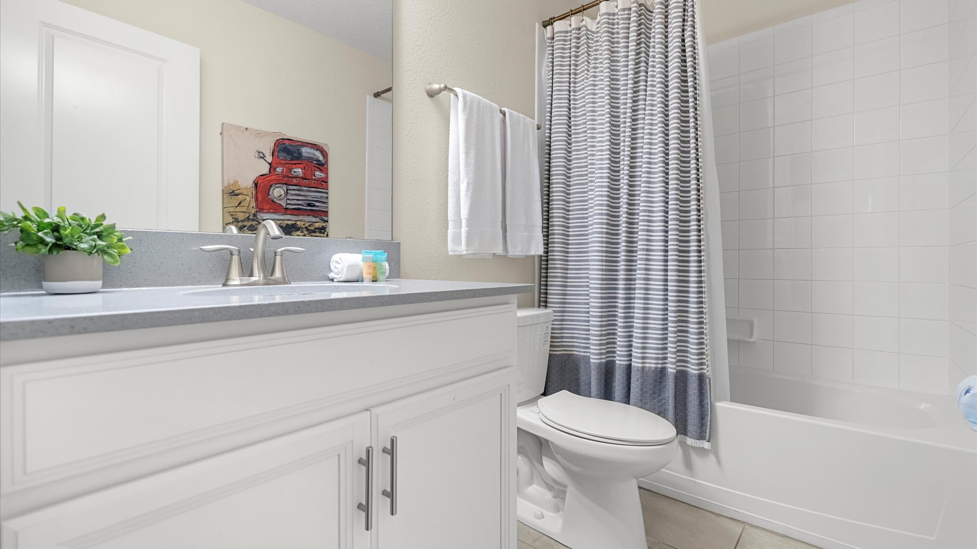 Bright bathroom featuring a white vanity with a grey countertop, a large mirror, and a white tiled shower/tub combo with a striped shower curtain. A red truck painting adds a pop of color.