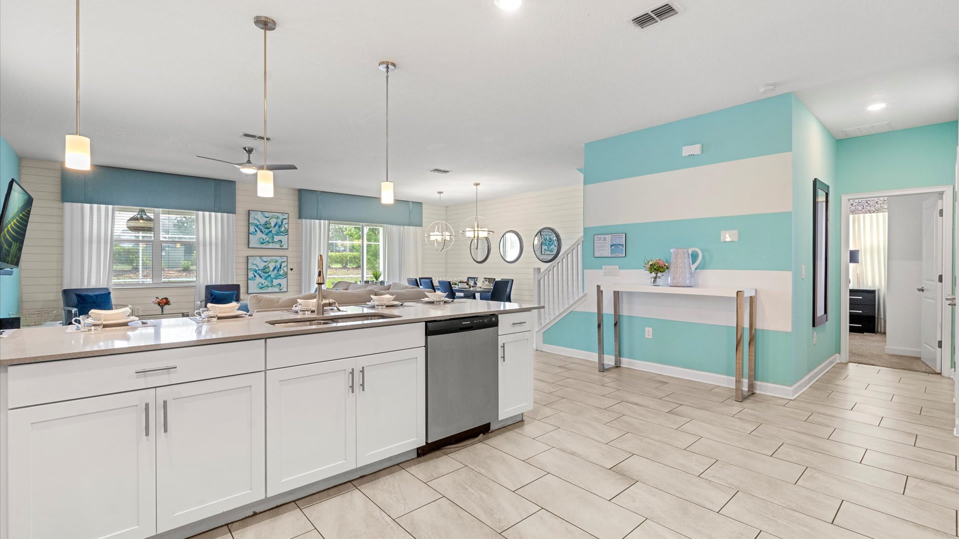 An open-concept living space features a modern kitchen island with white cabinetry and a dishwasher, overlooking a dining area with a large table and blue chairs, and a living room with a sofa, all under a white ceiling with recessed lighting and pendant lamps.