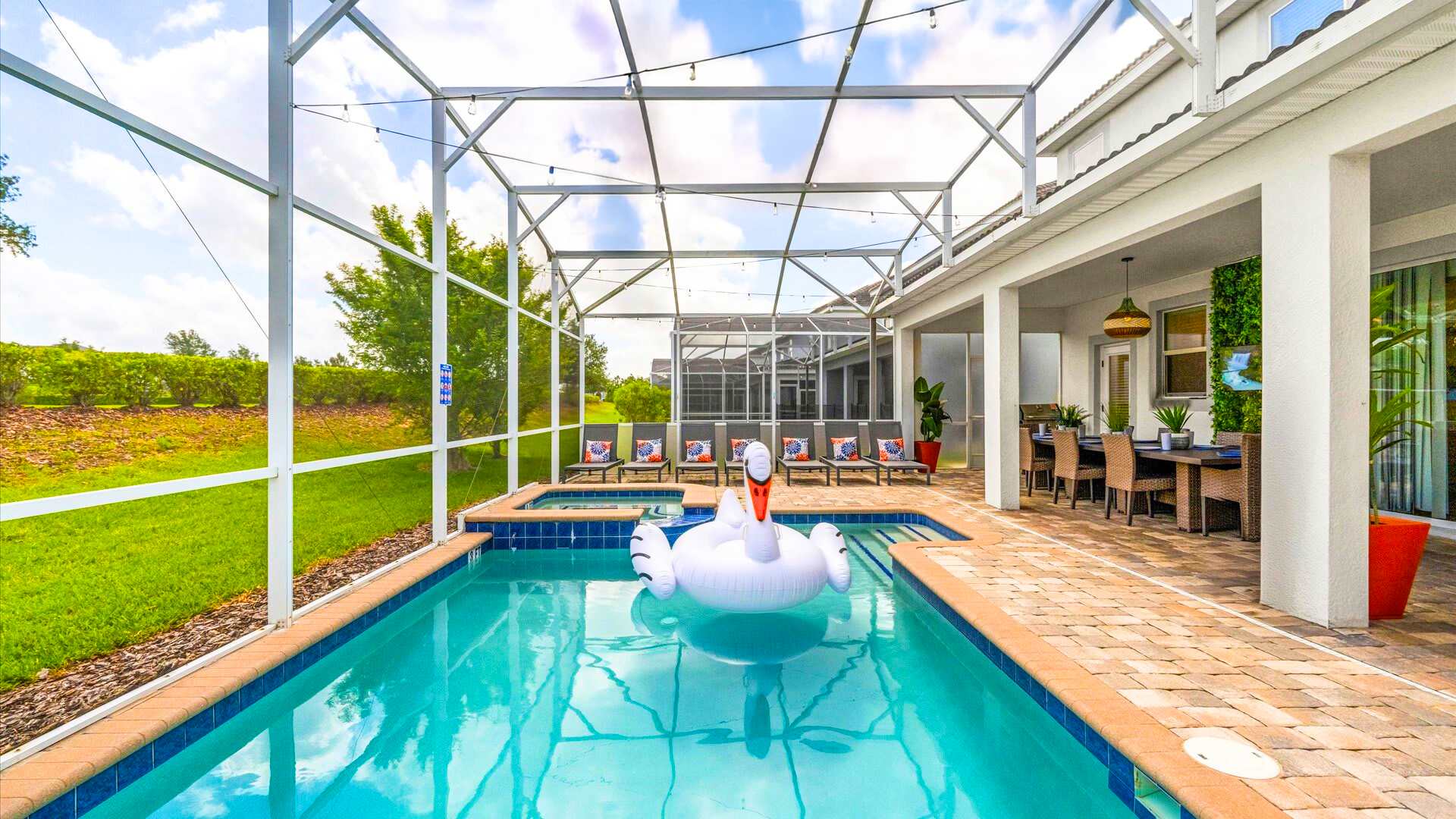 A wide shot of a screened-in private pool area with a hot tub, surrounded by brick pavers and several lounge chairs with colorful pillows. The pool overlooks a grassy backyard, and the covered lanai area is visible to the right.
