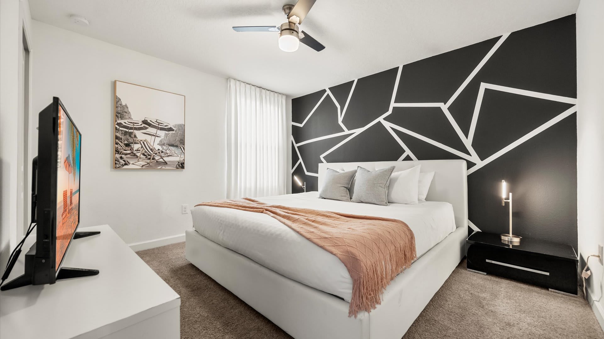 A modern bedroom featuring a white platform bed with a peach throw blanket, a large wall-mounted TV, and a striking black accent wall with white geometric patterns. A ceiling fan and two bedside lamps complete the contemporary look.