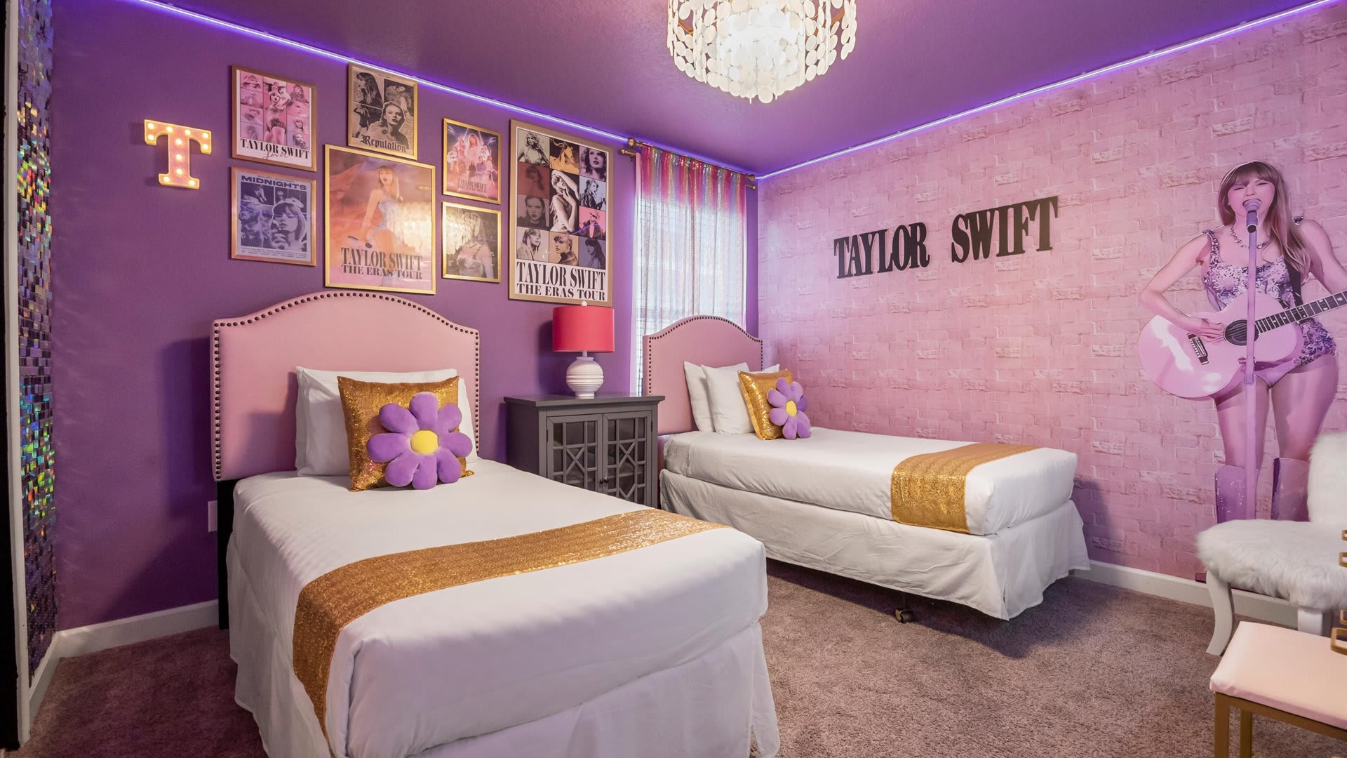 Themed bedroom with vibrant purple walls, two pink upholstered twin beds, and Taylor Swift posters adorning the wall. A unique chandelier hangs from the ceiling, and LED strip lighting adds ambiance.