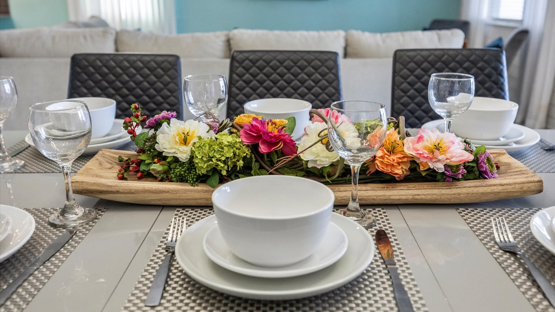 A detailed view of a dining table set with crisp white dinnerware and wine glasses, centered by a lush floral and greenery arrangement on a wooden tray. The modern setting includes black quilted dining chairs in the background.