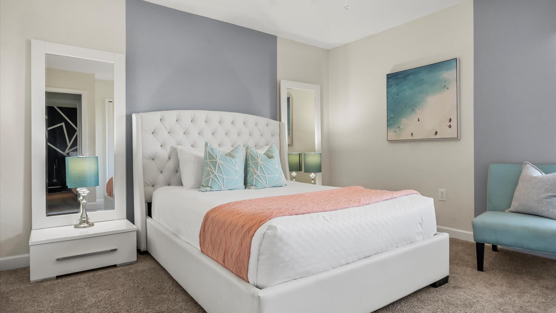 Elegant bedroom with a white tufted headboard bed, a grey accent wall, and two tall mirrors flanking the bed. Light blue and white throw pillows complement the room's serene color palette.